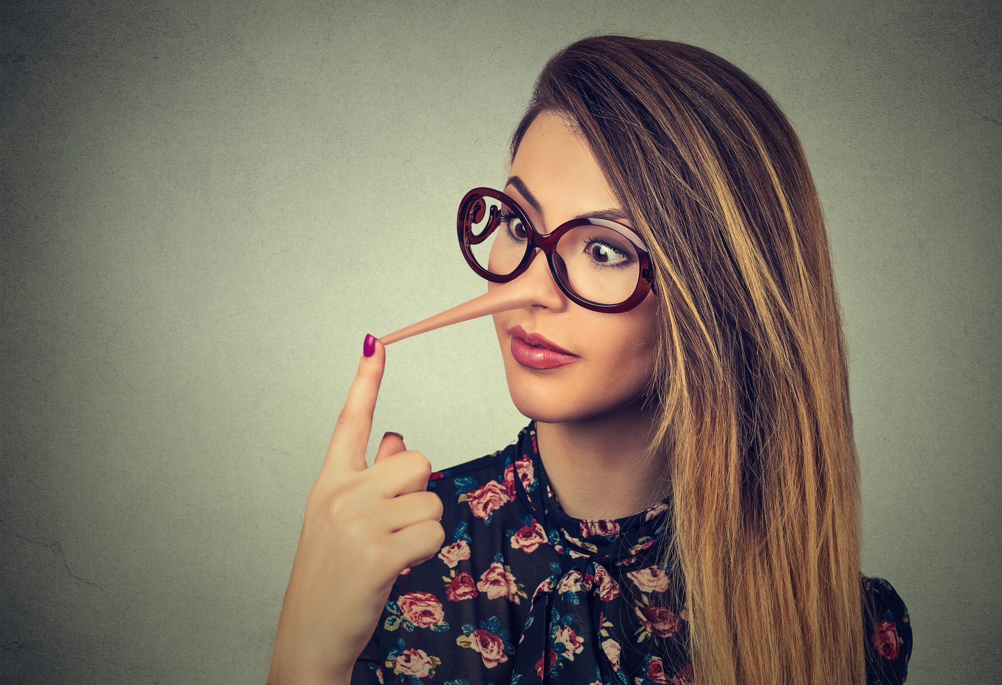 A woman with long hair and red lipstick wears large, round glasses and a floral top. She has a long Pinocchio-style nose and is touching the tip of it with her finger while looking surprised. She stands against a grey background.