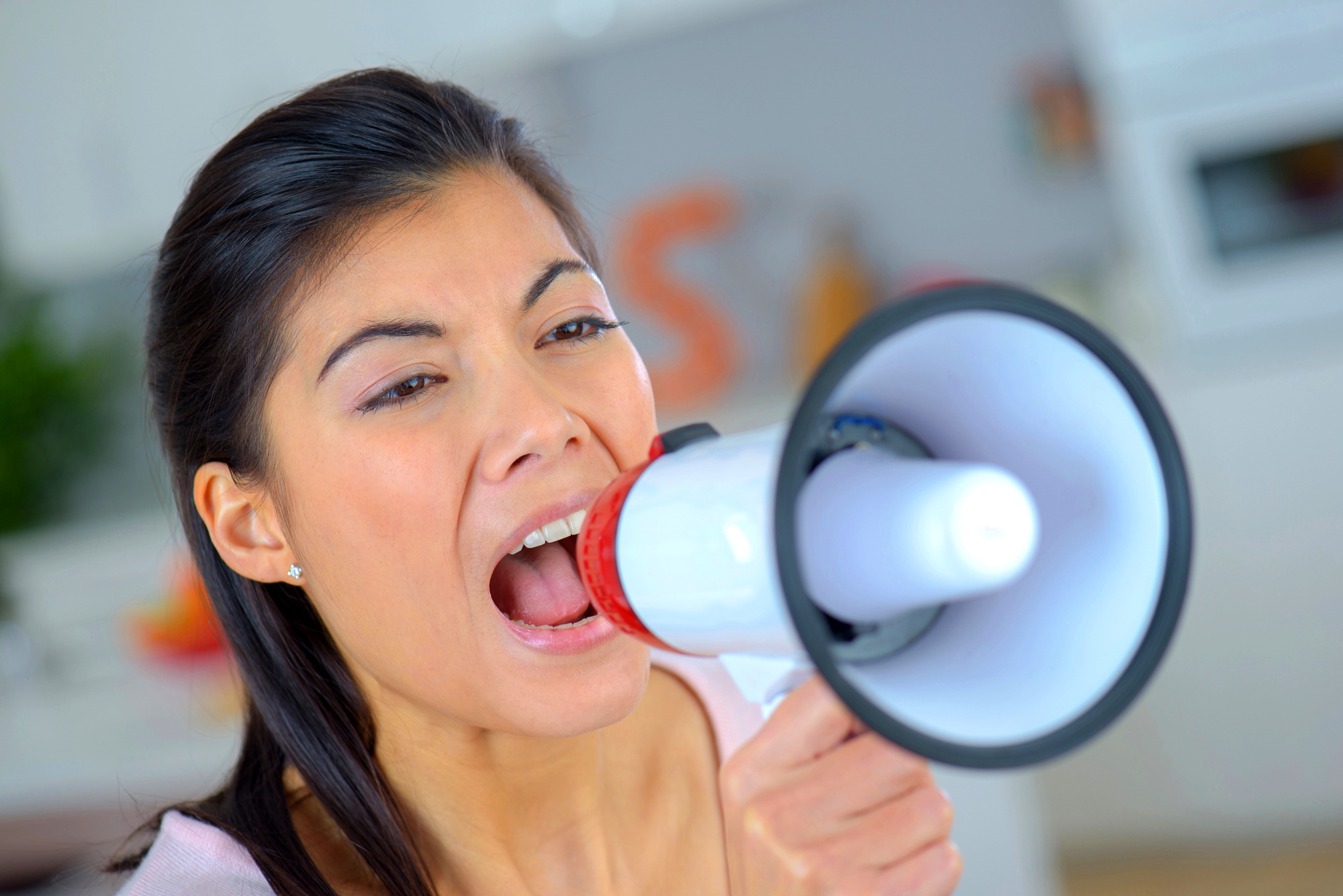A woman with dark hair tied back is shouting into a red and white megaphone. She is indoors, and the background is slightly blurred but appears to be a kitchen. Her expression suggests she is communicating something loudly.