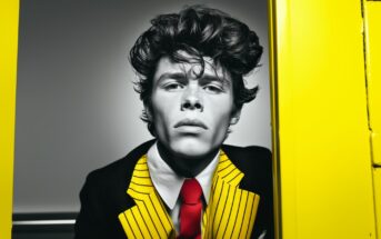 A young person with tousled hair wears a black jacket with bold yellow stripes, a white shirt, and a bright red tie, standing between two vibrant yellow panels. The image is in high contrast.