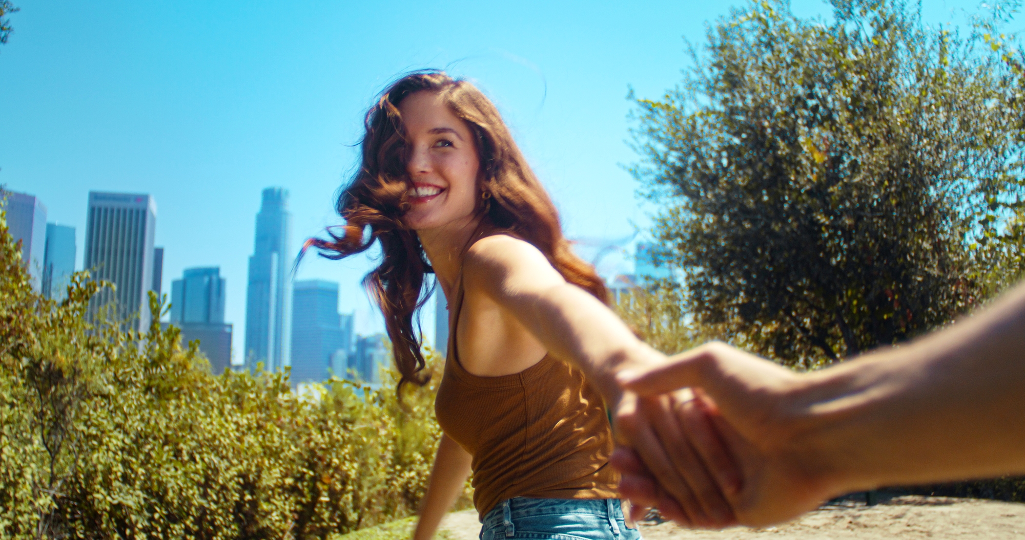 A smiling woman with long, wavy hair is leading someone by the hand through a scenic outdoor area with lush greenery. The skyline of a city with tall buildings can be seen in the background under a bright blue sky.