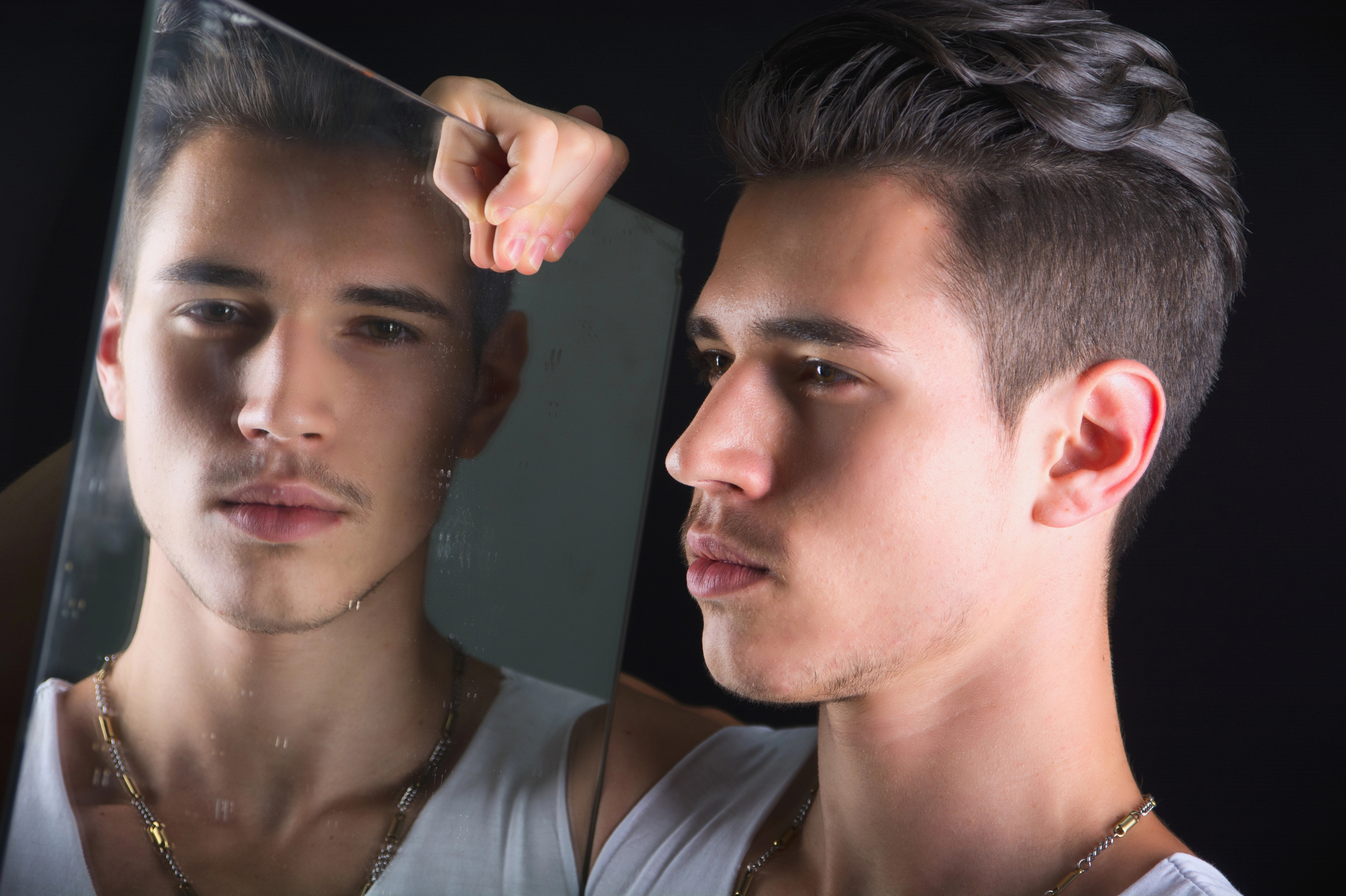 A young man with short dark hair and a light stubble is looking into a mirror, holding it with his right hand. His reflection in the mirror shows his pensive expression. He is wearing a white tank top and a gold chain necklace. The background is black.