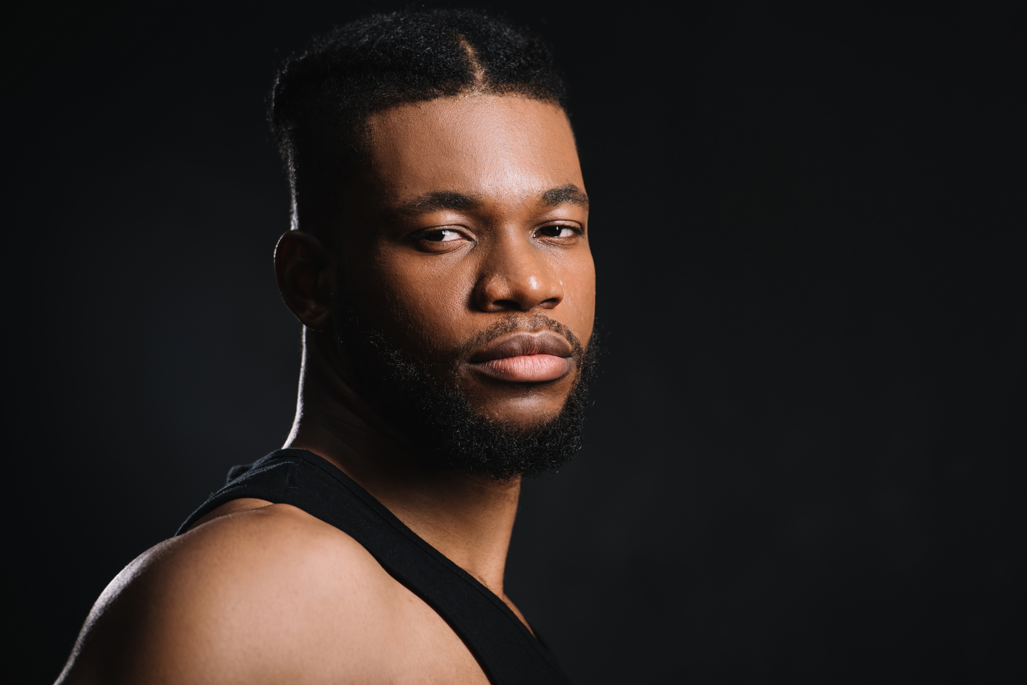 A man with a short beard and mustache gazes intently at the camera. He is wearing a sleeveless black top, and his hair is styled in a short, braided pattern. The background is a dark, neutral color, emphasizing his features.