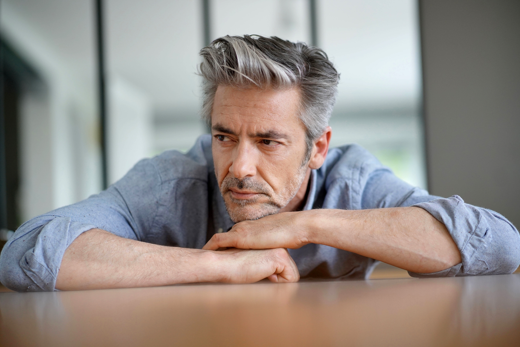 A middle-aged man with salt-and-pepper hair and a beard leans on a table, looking pensive. He is wearing a light blue button-up shirt and rests his chin on his forearm, appearing deep in thought. The background is out of focus, suggesting an indoor setting.