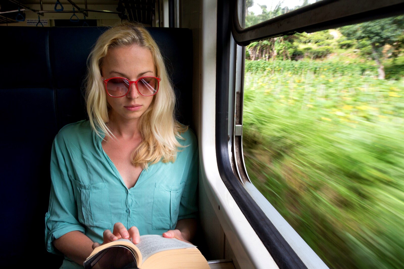A woman with blonde hair and red-framed glasses is seated by a window in a train, reading a book. She is wearing a light teal shirt. The view outside the window shows green, blurred scenery, indicating the train is moving.