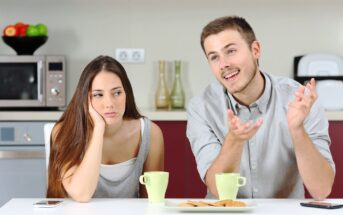 A man and woman sitting at a table with cups and cookies in front of them. The man is talking animatedly with hand gestures while the woman rests her head on her hand, appearing uninterested or bored. The setting is a kitchen with appliances and fruit in the background.