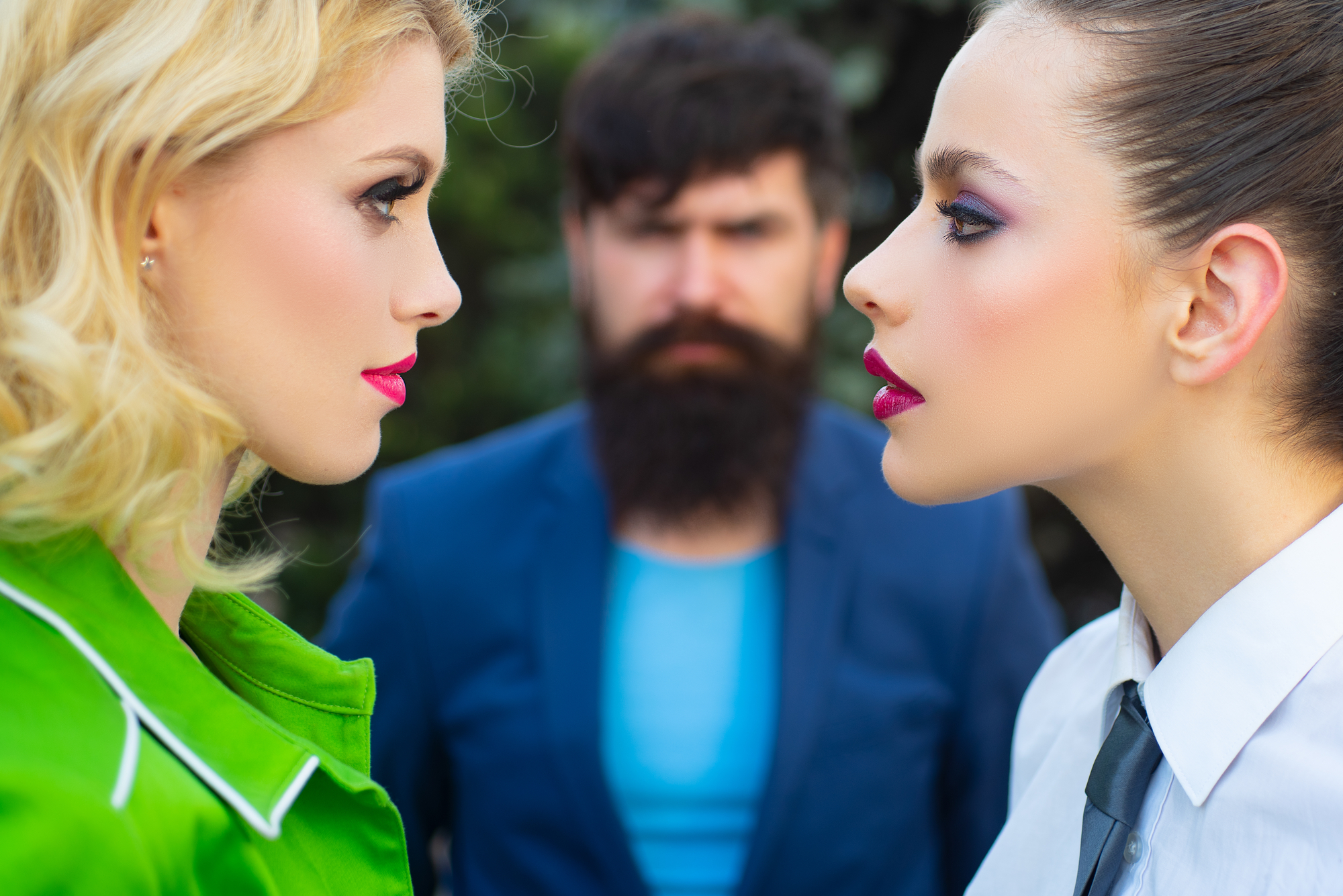 Two women face each other with serious expressions, one with blonde hair in a green jacket and the other with dark hair in white attire. A bearded man in a blue suit stands in the background, looking at them with a stern expression. The background is blurred.