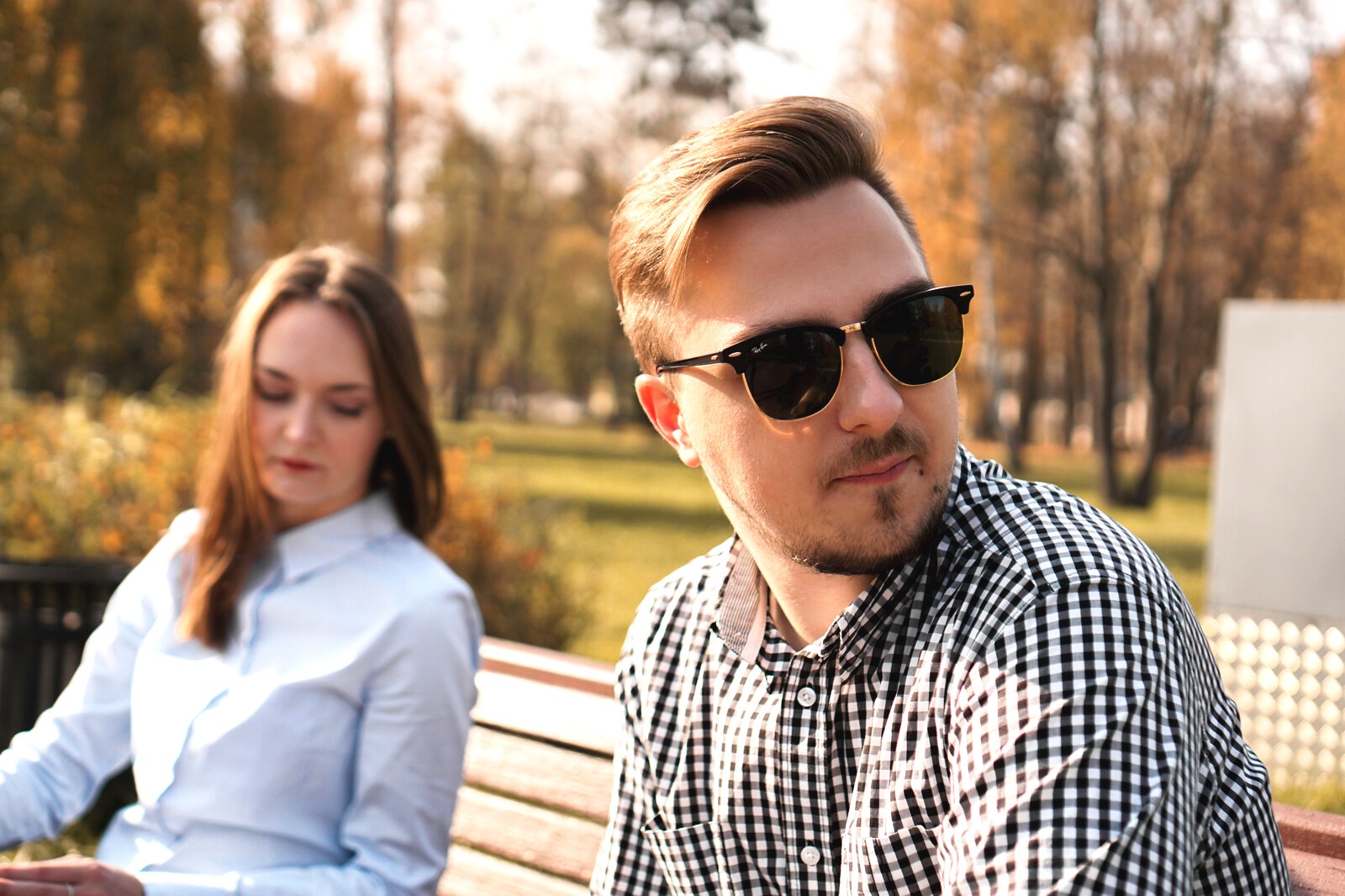A man wearing sunglasses and a checkered shirt looks to the side while sitting on a bench in a park. A woman with long brown hair, wearing a blue button-up shirt, sits next to him looking down. The background features trees with autumn leaves.