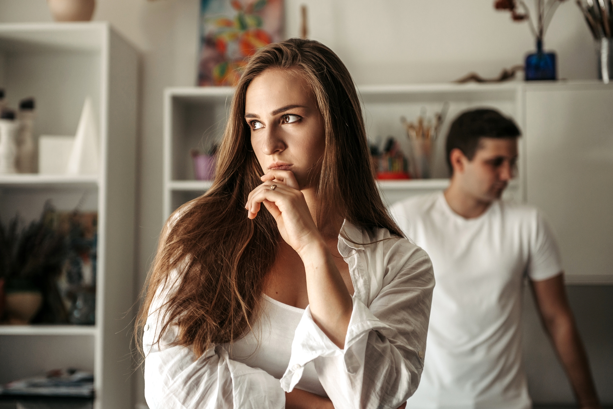 A woman with long brown hair and a thoughtful expression is wearing a white shirt and standing in an art studio. In the background, a man in a white t-shirt is slightly blurred, looking down. Shelves with art supplies and decorations are visible.