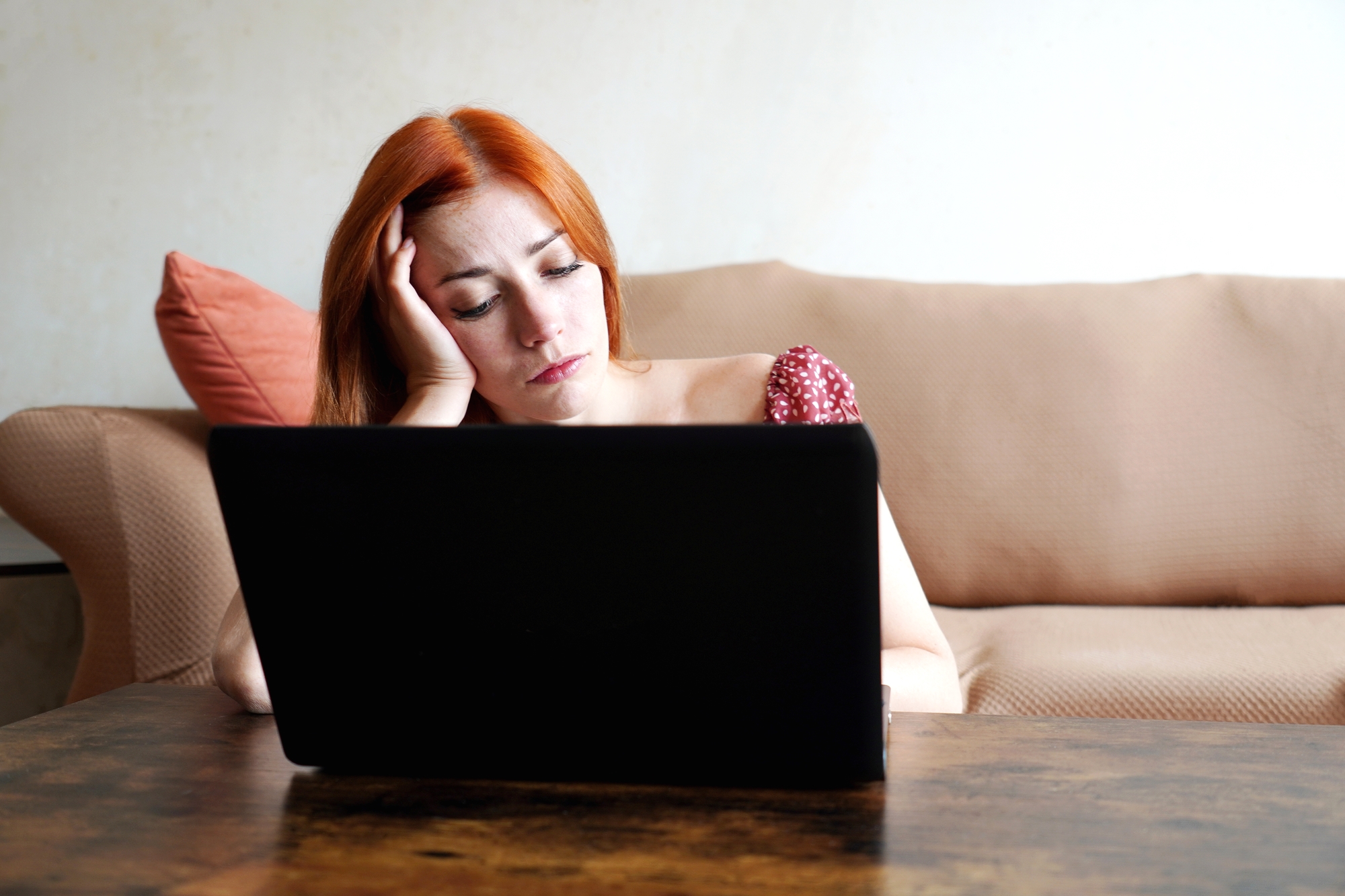 A woman with red hair wearing a red polka dot dress rests her head on her hand, looking tired or stressed, while sitting on a beige couch in front of an open laptop on a wooden table. A coral-colored pillow is in the background.
