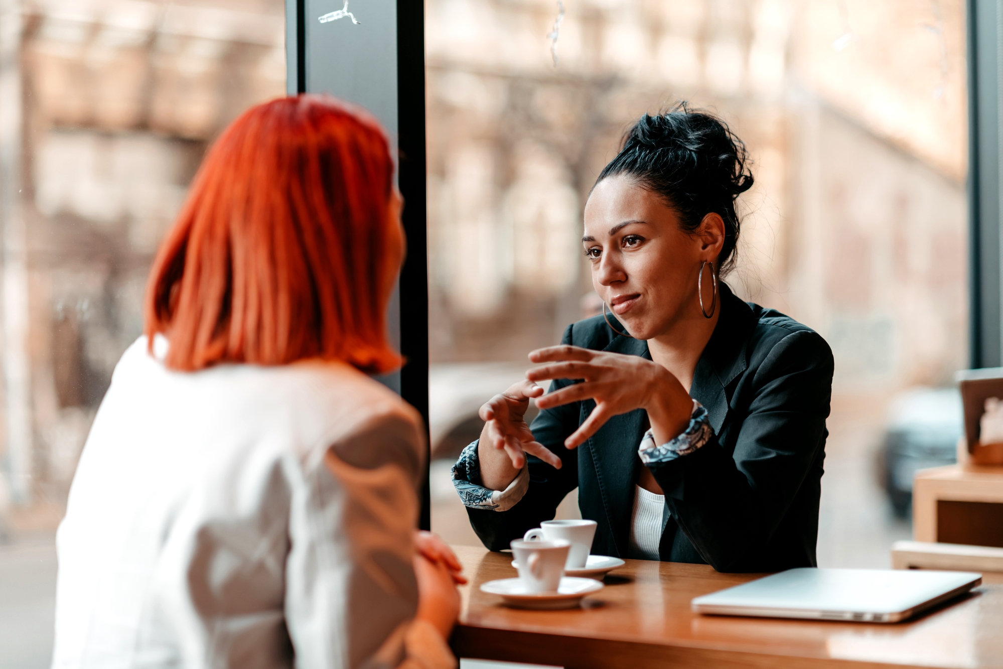 Two women sit at a wooden table in a cafe, engaging in an animated conversation. One woman has red hair and wears a white blazer, while the other has dark hair tied up and wears a black blazer. Each has a small cup of coffee in front of them beside a laptop.