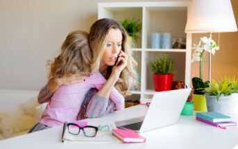 A woman sits at a desk, talking on the phone with a concerned expression, while holding a child in her lap. The desk has a laptop, notebook, glasses, and various plants. The room is brightly lit with a table lamp and decorated with plants and colorful items.