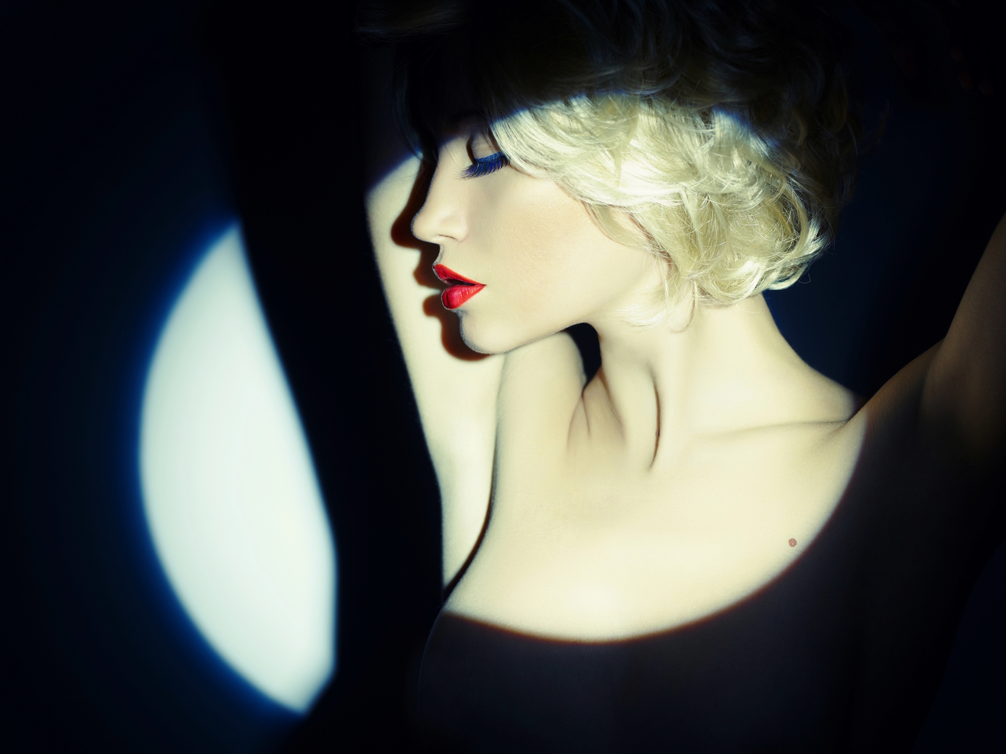 A woman with short blonde hair and red lipstick poses with her arms raised above her head. She is illuminated by a focused, blue-tinted spotlight, creating dramatic shadows against a dark background. Her eyes are closed, and she conveys a feeling of poise and elegance.