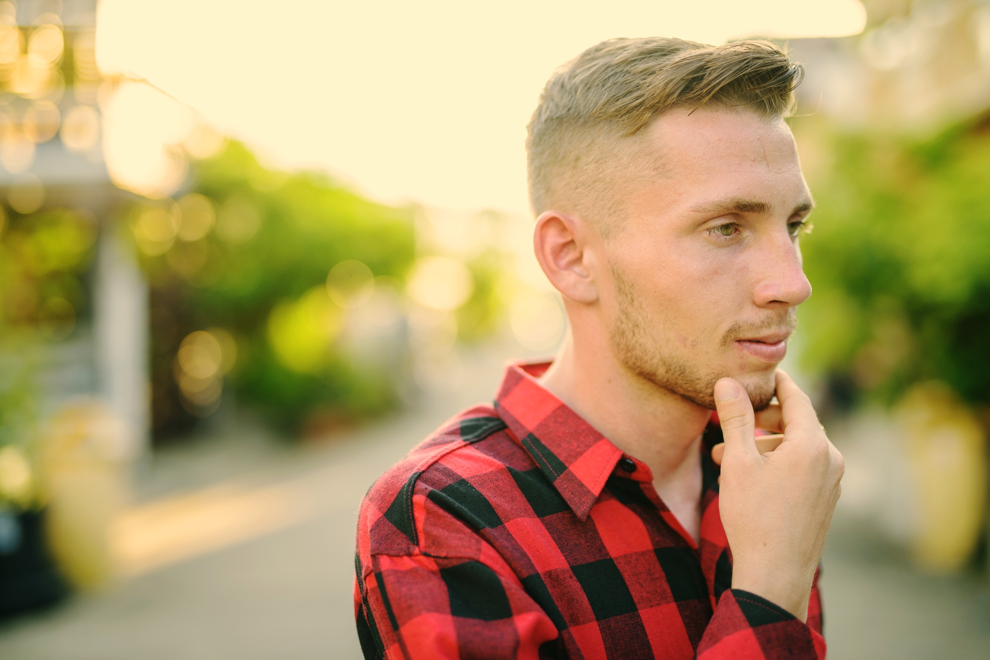 A young man with blonde hair and a beard, wearing a red and black checkered shirt, stands outdoors on a sunny day. He is looking to the side with one hand thoughtfully touching his chin. The background is out of focus, featuring greenery and a blurred pathway.