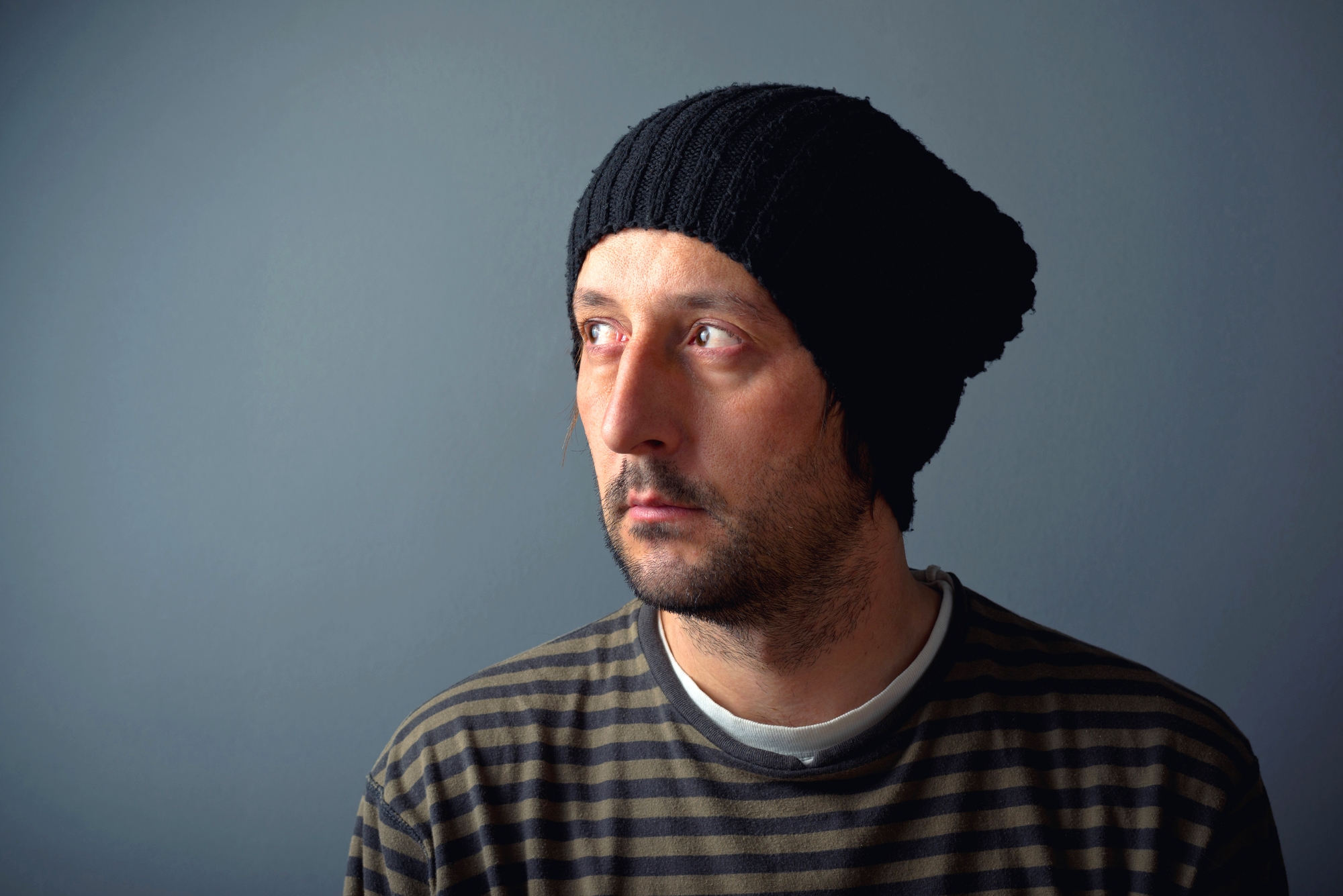A man with a tired and contemplative expression looks toward the upper left. He has red, irritated eyes and wears a black knit beanie and a dark striped shirt. The background is a solid, muted gray.