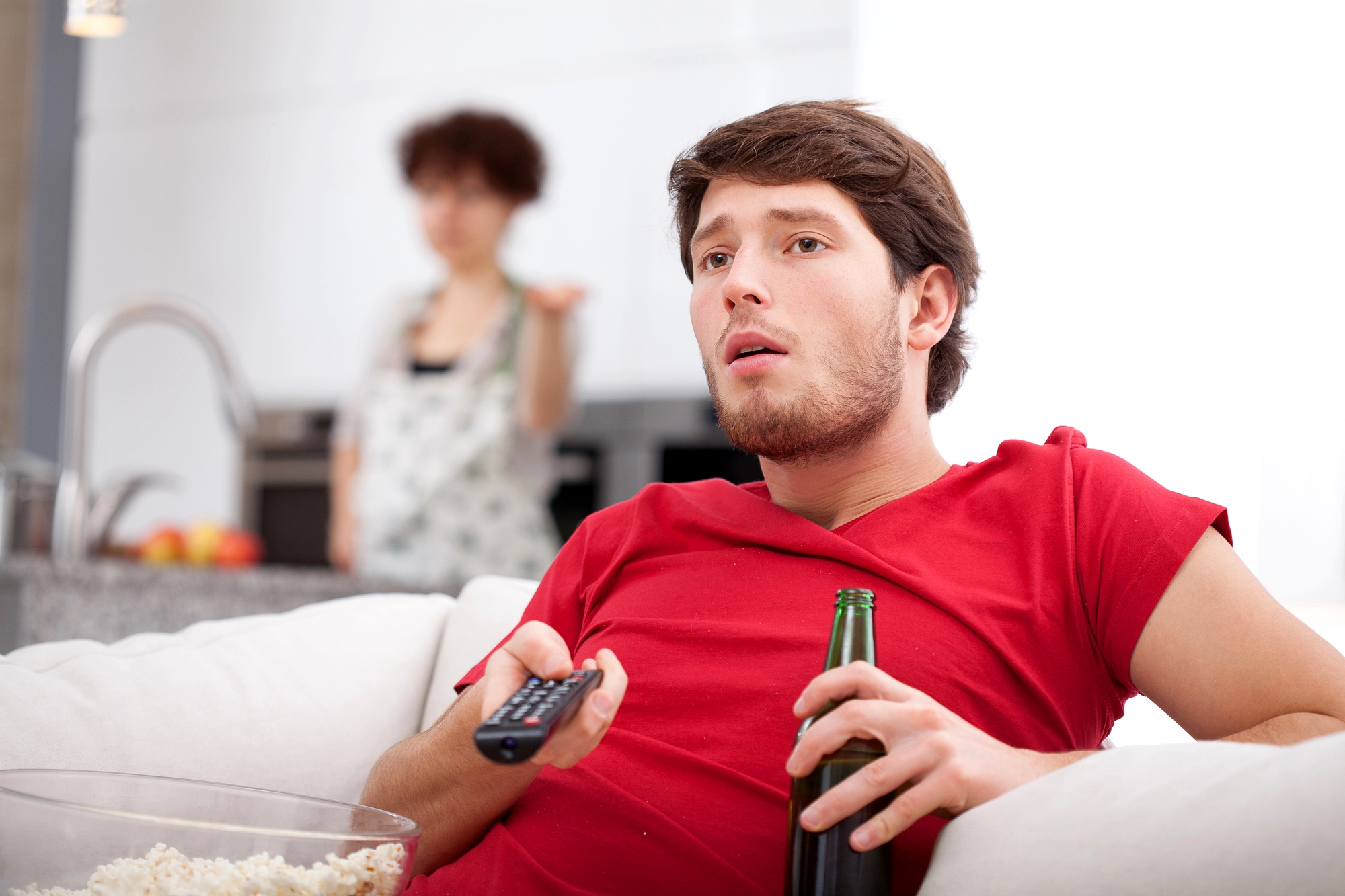 A man in a red shirt sits on a couch holding a TV remote and a beer bottle, looking intently at the television. A woman in the background, standing in the kitchen, appears to be talking or gesturing towards the man. A bowl of popcorn is in the foreground.