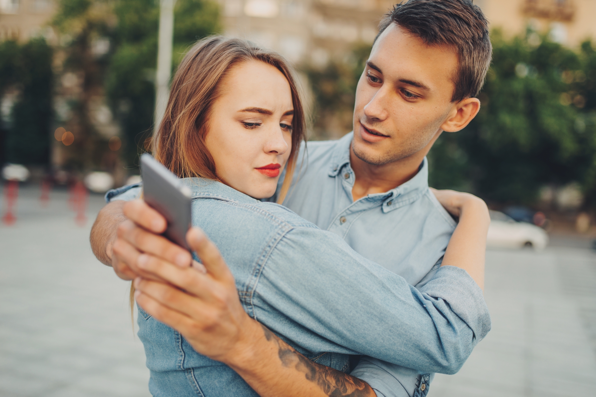A young couple wearing denim shirts stands close together outdoors. The woman holds out a smartphone, looking concerned, while the man has one arm around her and points at the phone screen with a focused expression. Trees and buildings can be seen in the background.