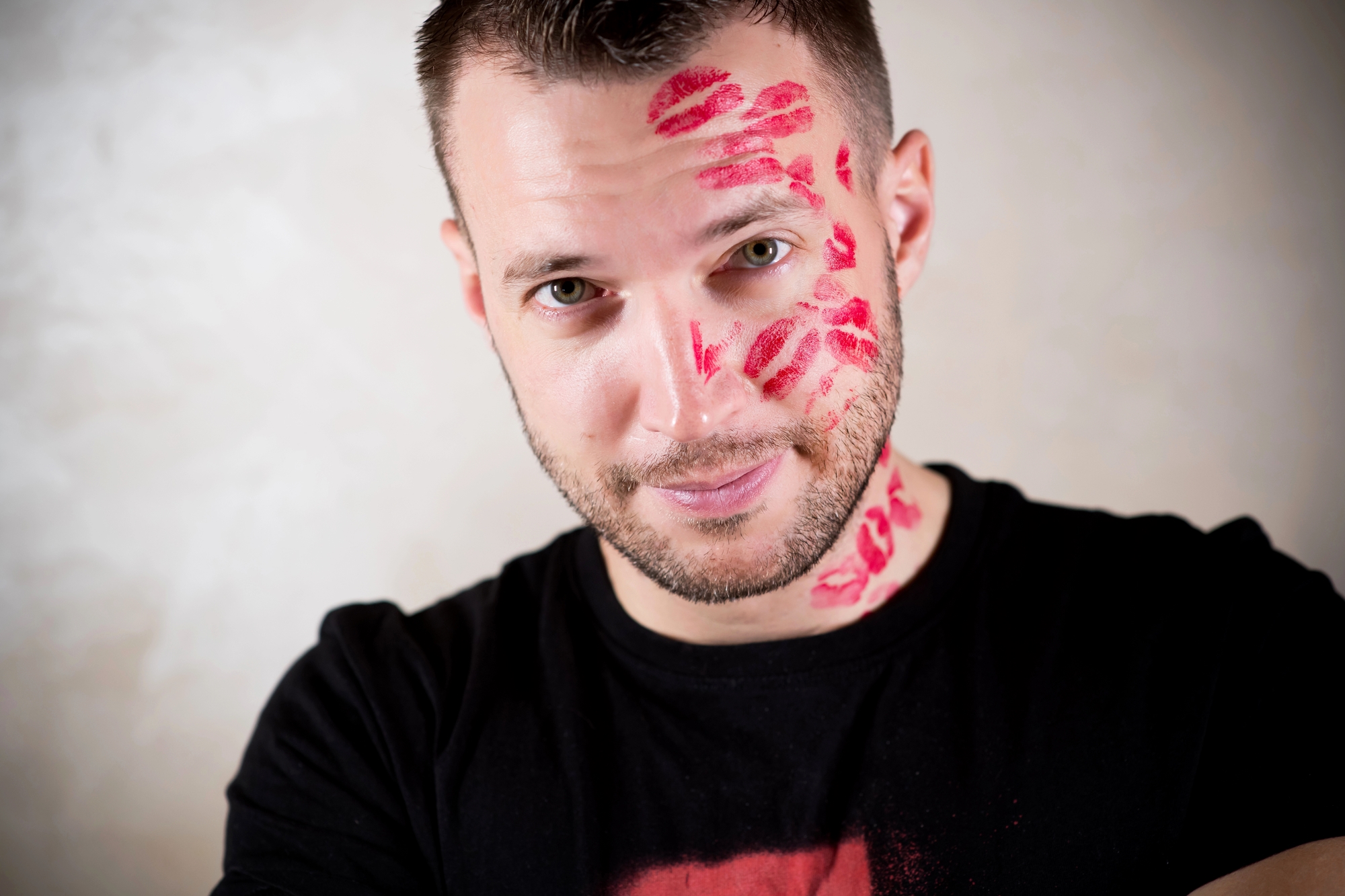 A man with short hair and a beard is smiling at the camera. His face and neck are covered in numerous red lipstick kiss marks. He is wearing a black t-shirt and has a playful expression. The background is a plain, light-colored wall.