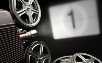 A close-up of an old-fashioned film projector with several reels of film shown on the left side. The background is dark, with a blurred countdown screen displaying the number "1" in white, indicating the start of a film.