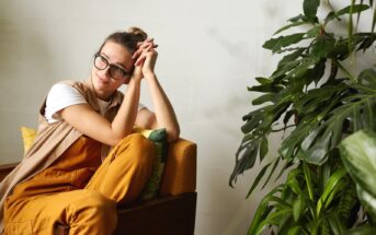 A person wearing glasses and a yellow outfit sits on a couch, leaning on their hands and looking thoughtfully to the side. The background features a white wall and a large green plant.