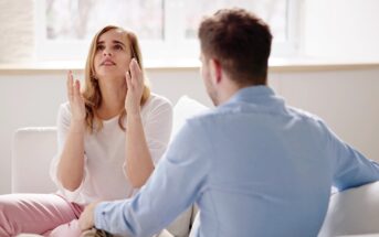 A woman with long brown hair, wearing a white top and pink pants, sits on a couch gesturing with her hands up, looking distressed. She is talking to a man with short hair wearing a light blue shirt and beige pants, who is facing her with his back to the camera.