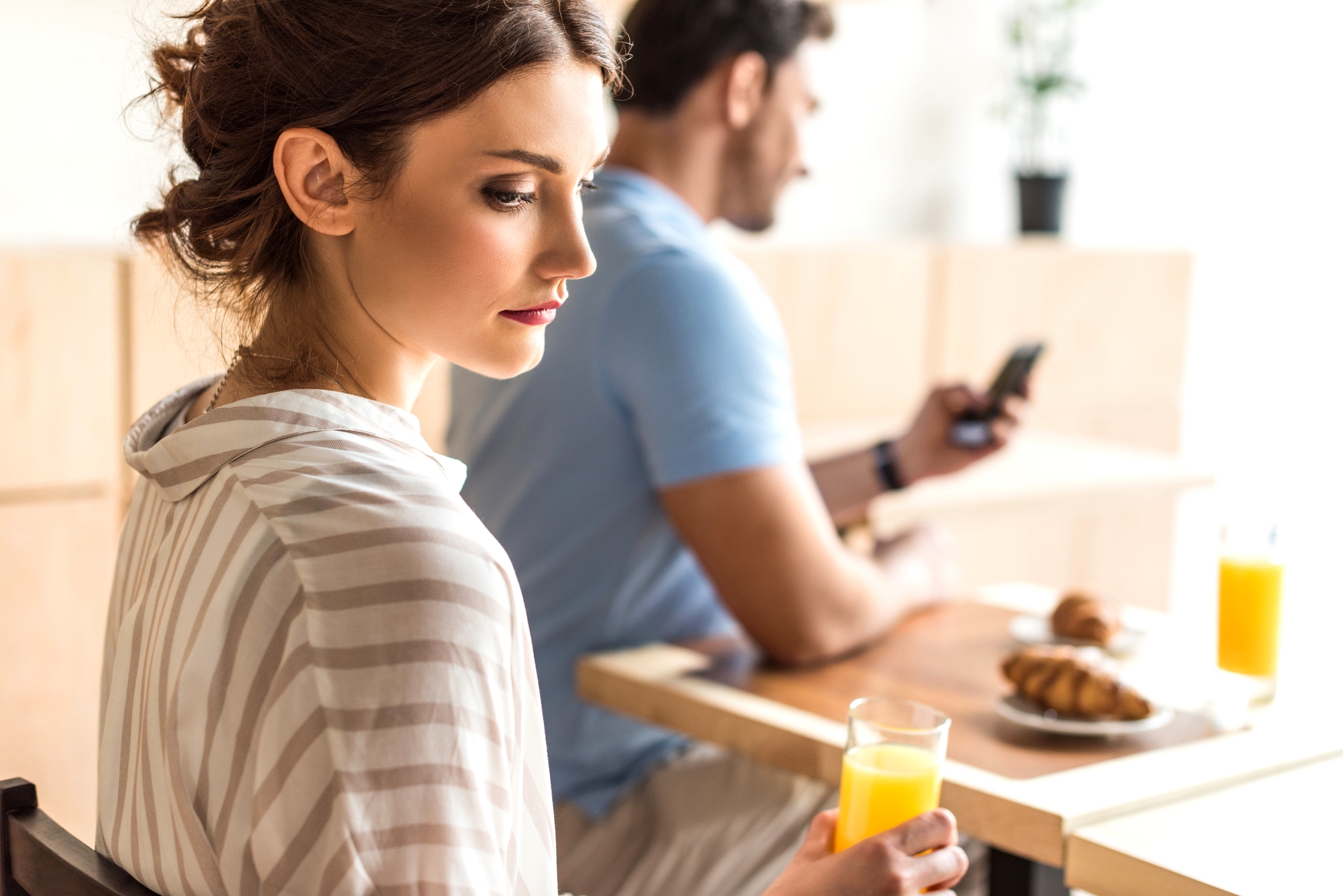 A woman with brown hair wearing a striped shirt holds a glass of orange juice while looking to the side, sitting at a table. In the background, a man with dark hair and a blue shirt looks at his phone. Croissants and orange juice are on the table.