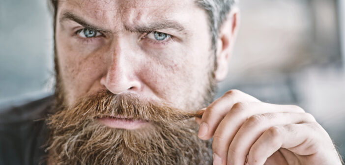 A bearded man with blue eyes intensely gazes into the camera while twirling a strand of his mustache with his right hand. The background is blurred, focusing the attention on his expressive face.