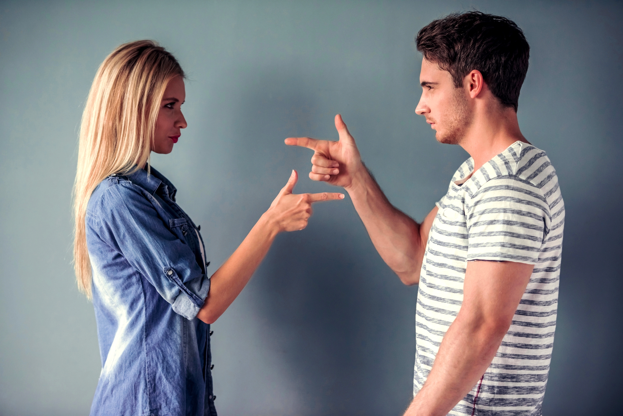 A woman and a man are standing facing each other against a gray background. Both are making playful hand gestures, mimicking guns pointed at each other. The woman has long blonde hair and is wearing a denim shirt, while the man has short brown hair and is wearing a striped shirt.