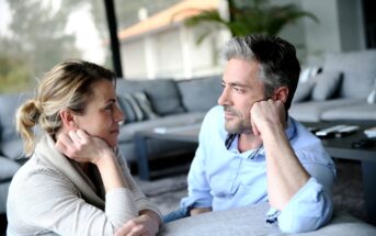 A man and a woman sit facing each other, resting their heads on their hands in a cozy and modern living room with gray furniture and large windows. They are engaged in an intimate and thoughtful conversation. A lush plant is visible in the background.