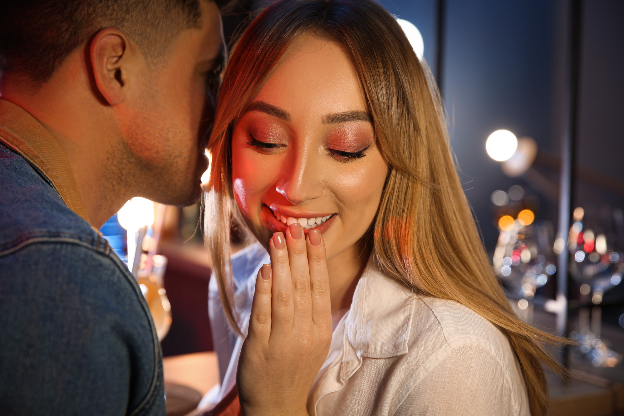 A person is whispering into the ear of a smiling woman with long hair. The woman, covering her mouth with her hand, looks amused. The background is softly lit with various lights and blurred details, suggesting an evening or indoor setting.