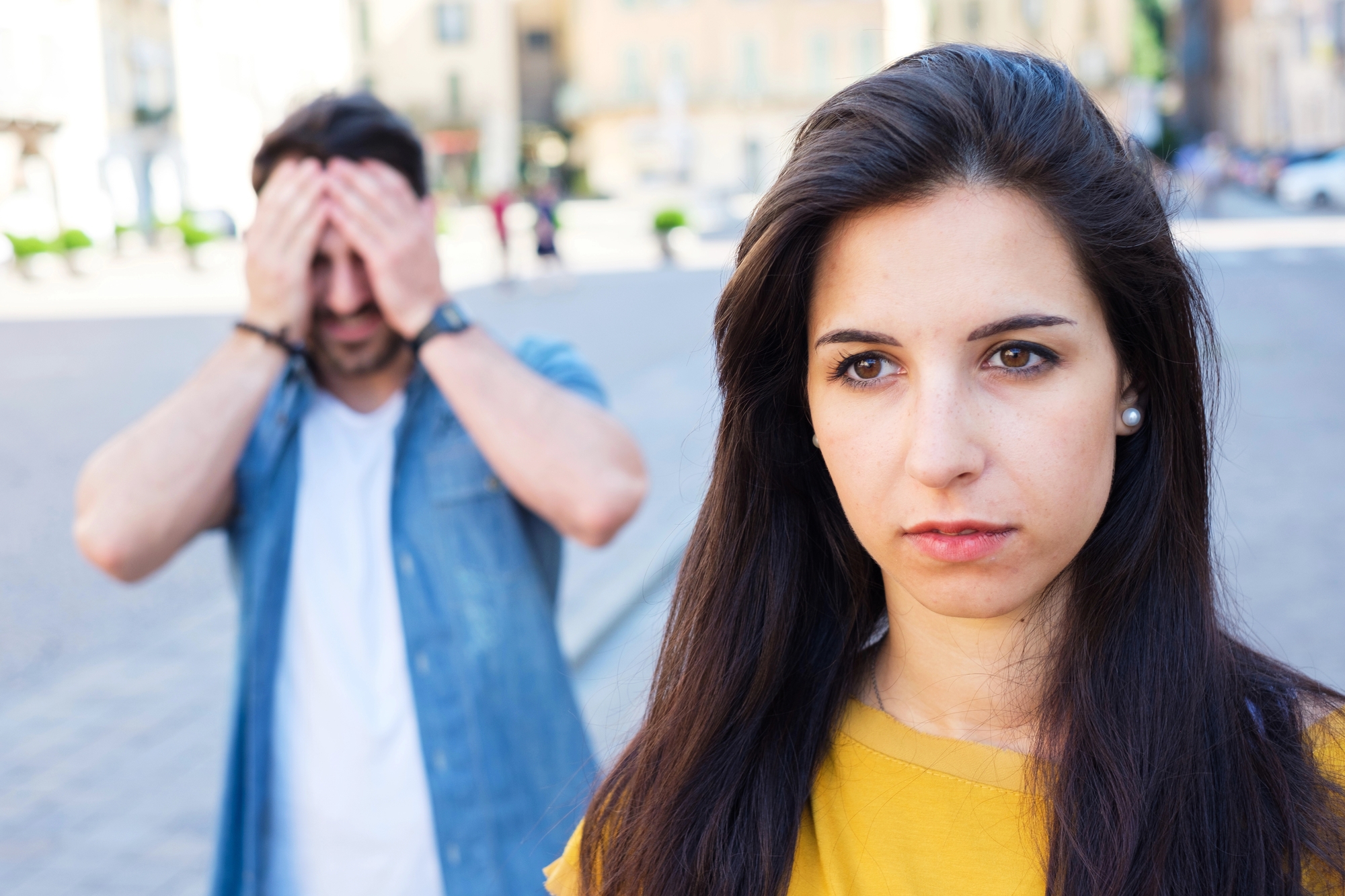 A woman with long dark hair looks serious and distant in the foreground. In the background, a man in a denim shirt covers his face with his hands, standing on a city street with blurred buildings and people. The scene suggests tension between them.
