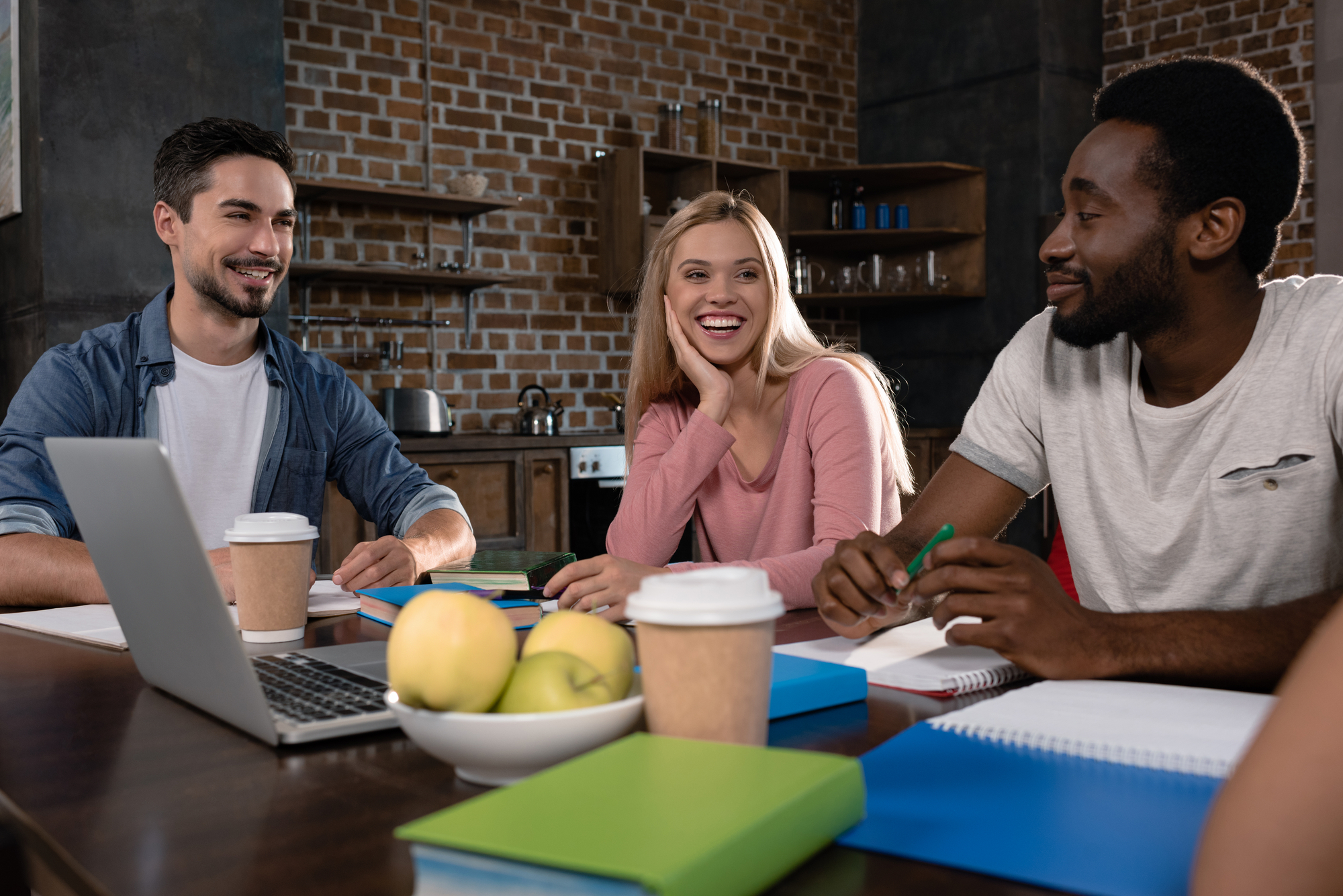 Three young adults are sitting around a table in a cozy, brick-walled room, smiling and chatting. They have books, a laptop, and coffee cups on the table, with a bowl of fruit. The atmosphere is relaxed and conversational.