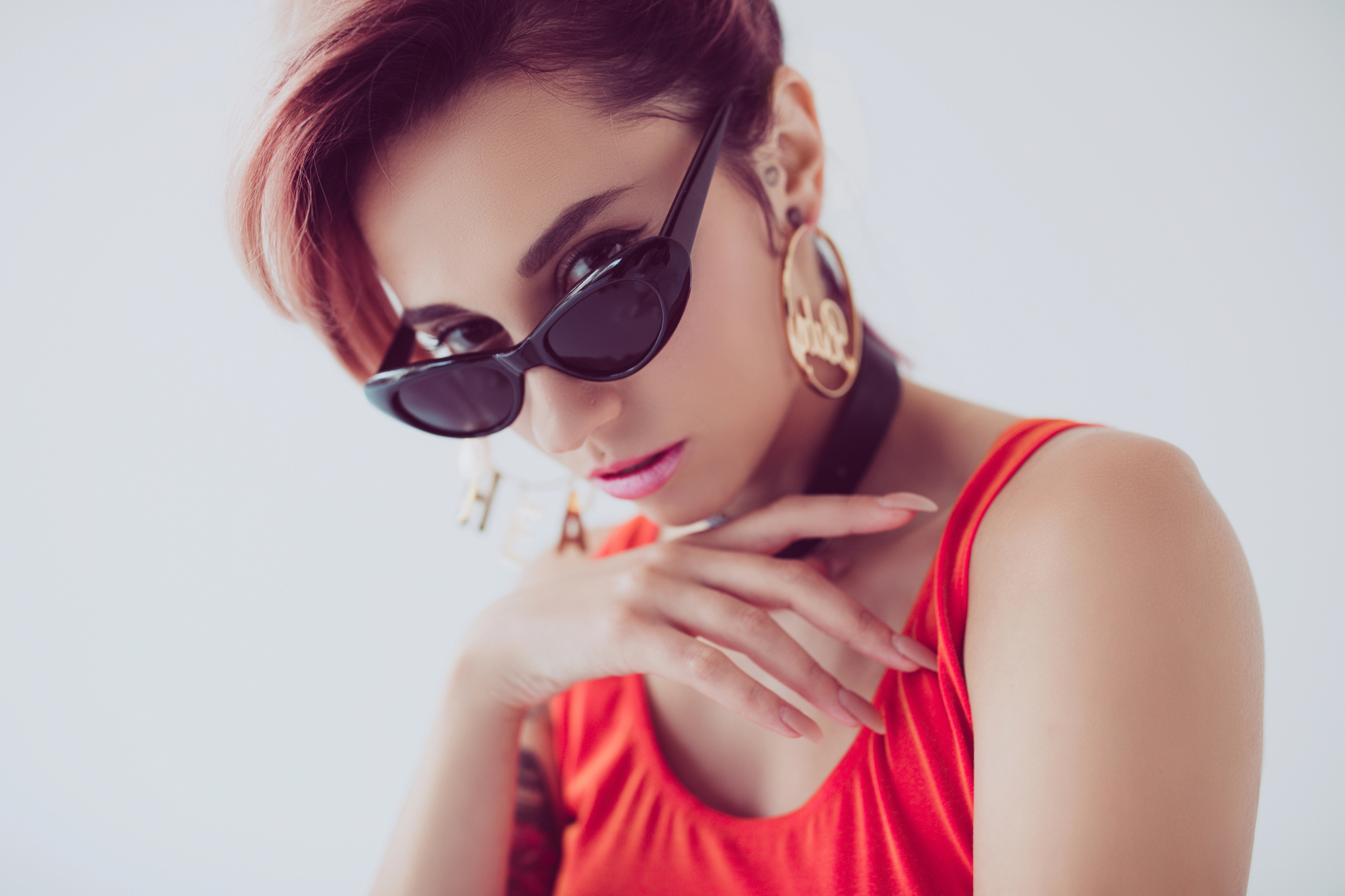 A woman with short, red hair wearing cat-eye sunglasses, large hoop earrings, and a vibrant red top poses with her hand near her face against a plain white background. She has a confident, stylish look.