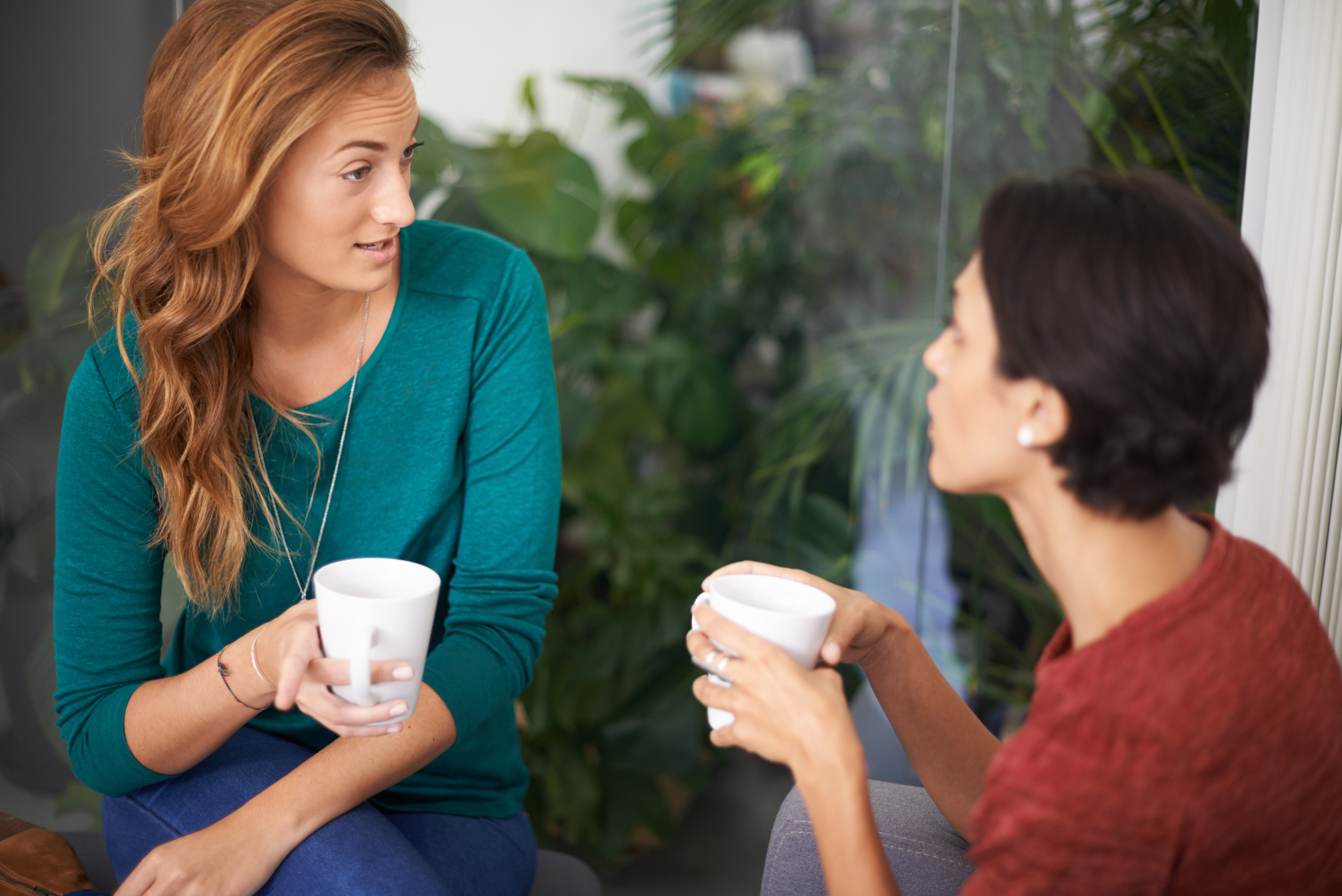 Two women are sitting and holding white mugs while engaging in conversation. The woman on the left has long, wavy hair and wears a teal top, while the woman on the right has short hair and wears a reddish-brown top. Lush green plants are visible in the background.