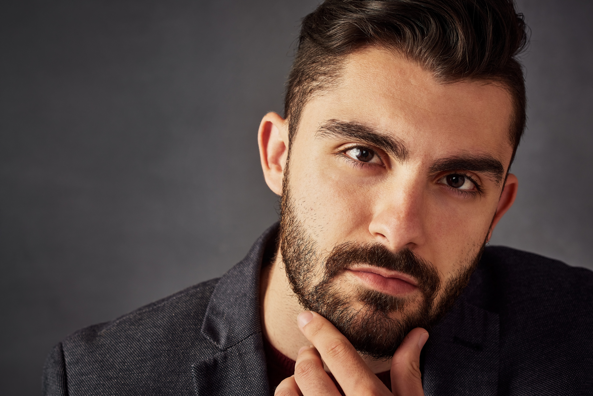 Close-up portrait of a man with a well-groomed beard and dark hair, wearing a dark blazer. He is looking directly at the camera with a thoughtful expression, his hand resting on his chin against a dark, neutral background.