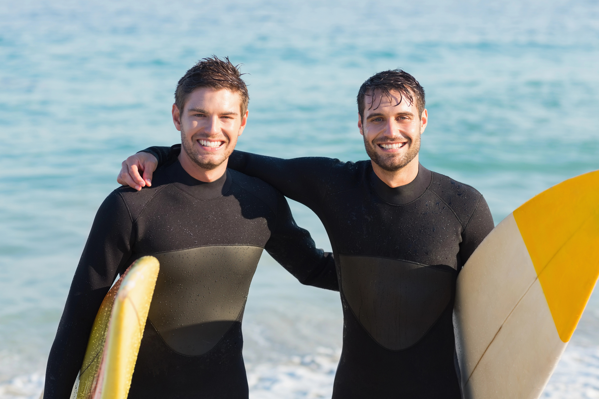 Two men wearing wetsuits and holding surfboards stand side by side on a beach, smiling at the camera. The ocean waves in the background indicate an ideal surfing location.