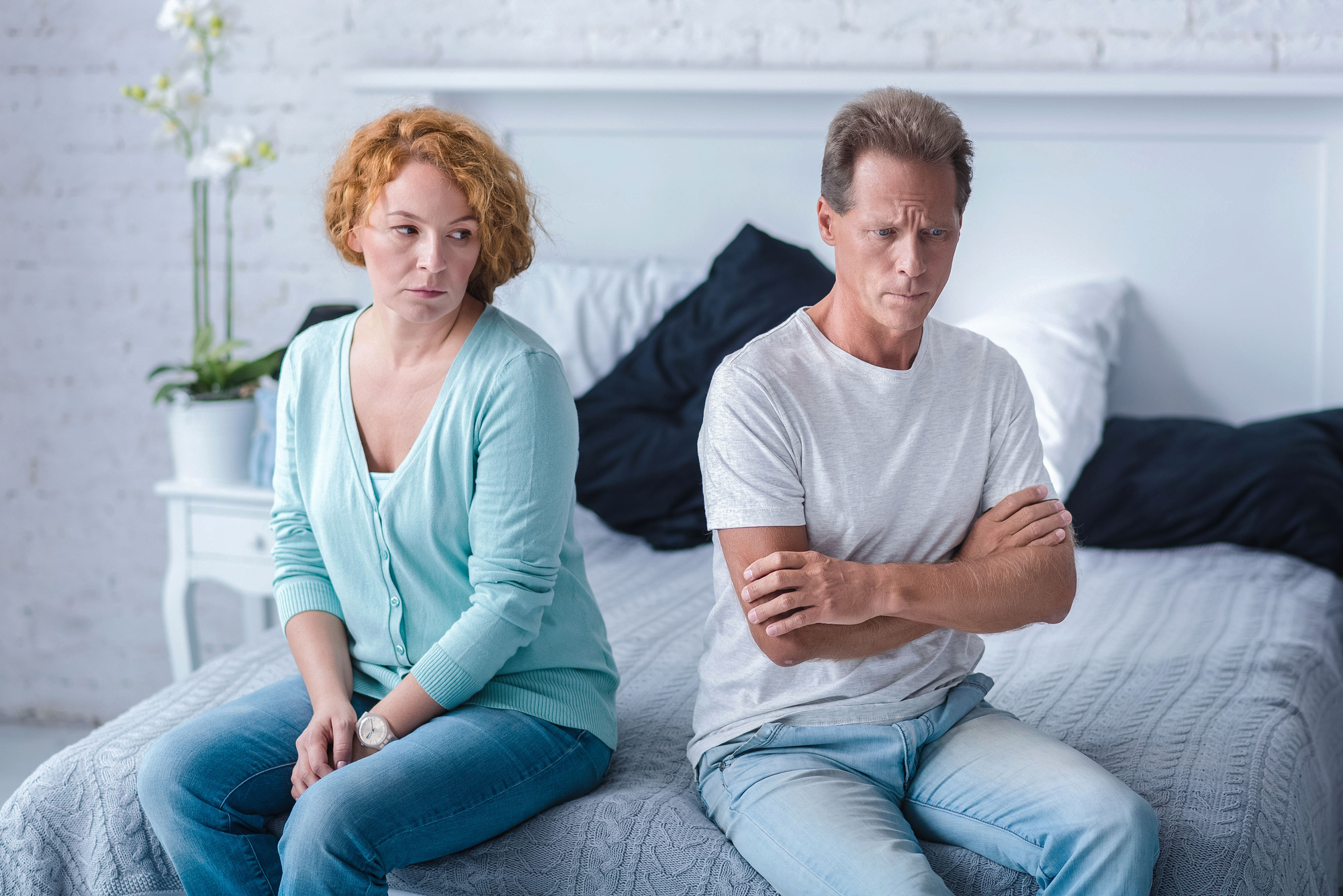 A woman with curly red hair and a light blue cardigan sits on a bed, looking to her left with a concerned expression. A man with short hair and a white t-shirt sits next to her but faces the opposite direction, appearing upset with his arms crossed.