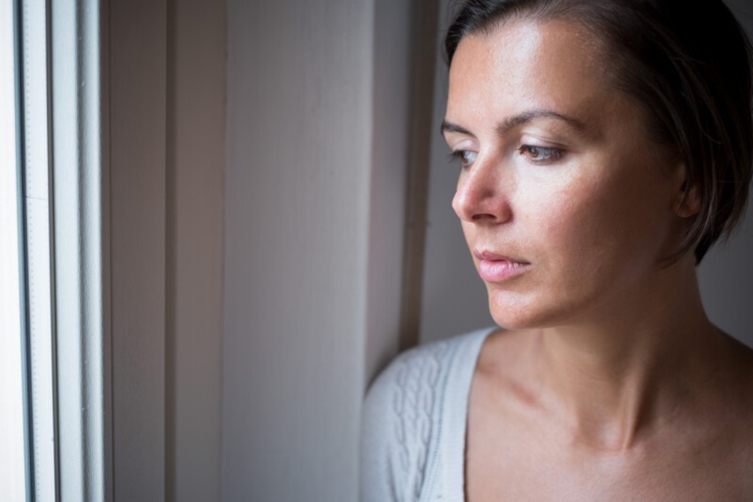 A contemplative woman with short brown hair is gazing out of a window. She appears thoughtful, with a neutral expression on her face. She is dressed in a light-colored top and is bathed in soft, natural light filtering through the window.