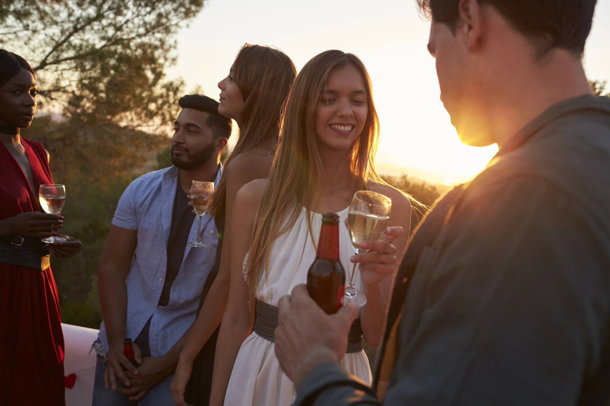 A group of young adults is enjoying an outdoor gathering at sunset, holding drinks and smiling. The warm light of the setting sun illuminates their faces as they converse. Trees and a scenic landscape are visible in the background.
