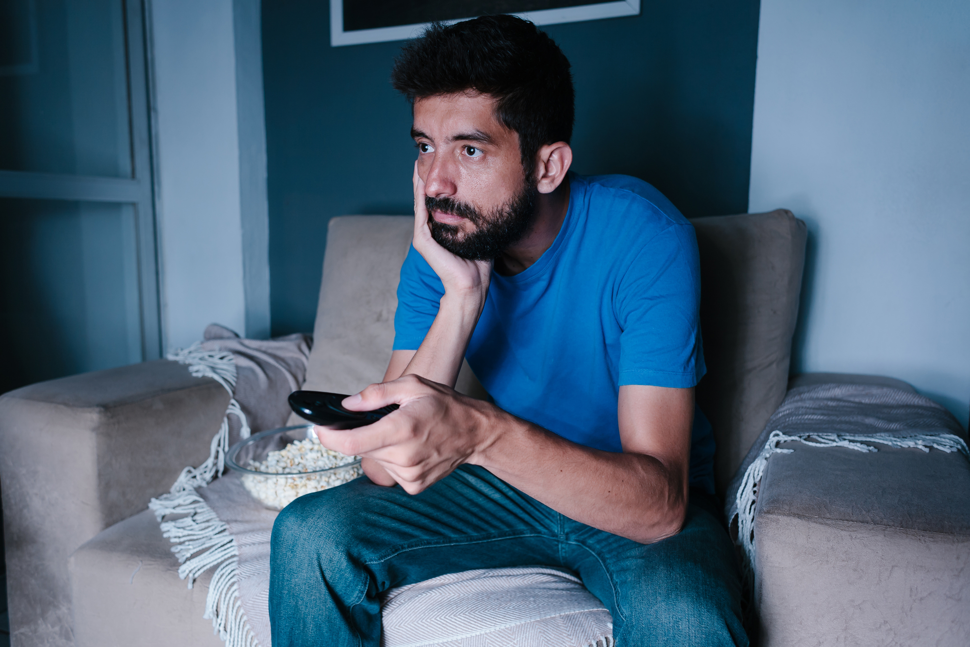 A man with short dark hair and a beard sits on a beige armchair, holding a TV remote in one hand while resting his head on the other. He seems uninterested or bored. A bowl of popcorn is placed on the arm of the chair beside him.