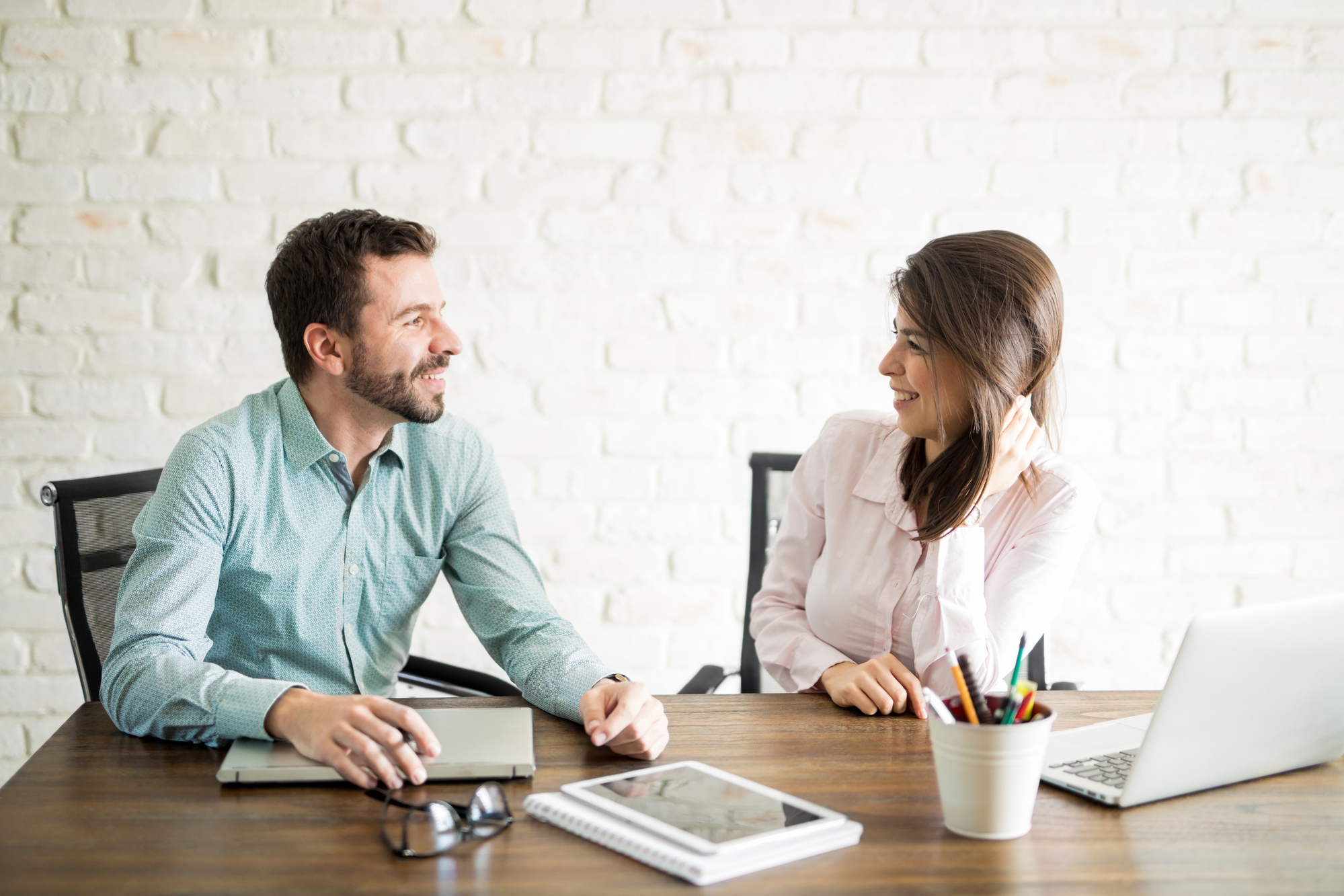 A man and a woman are sitting at a wooden table in a modern office setting. They are smiling and engaged in a conversation. On the table are a laptop, a tablet, a notebook, glasses, and a cup holding various pens and pencils.
