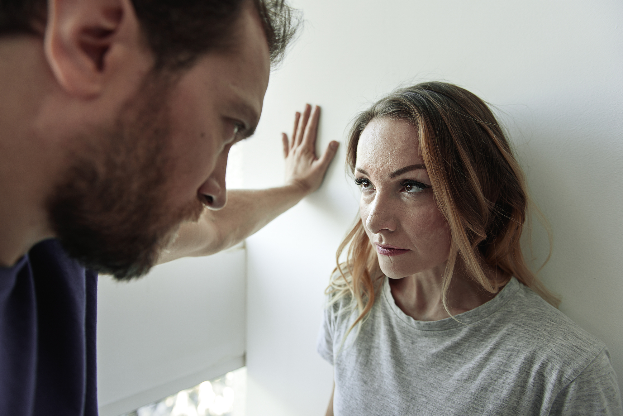 A man stands close to a woman, with his arm raised and hand against the wall, looking down at her. The woman looks up at him with a serious expression. The scene suggests a tense or confrontational moment.
