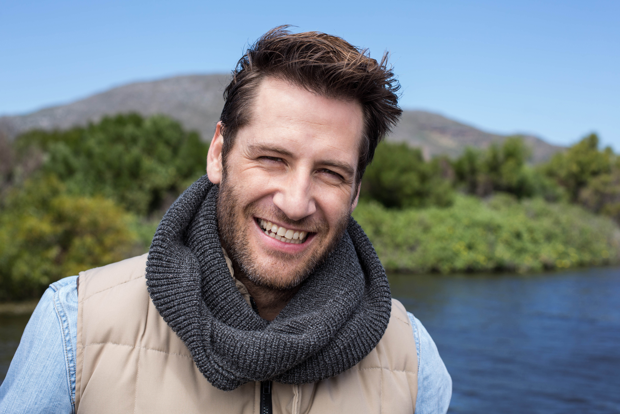 A man with short brown hair and a beard smiles at the camera. He is wearing a beige jacket and a thick, gray scarf. Behind him is a scenic outdoor background featuring a body of water, lush greenery, and distant hills under a clear blue sky.