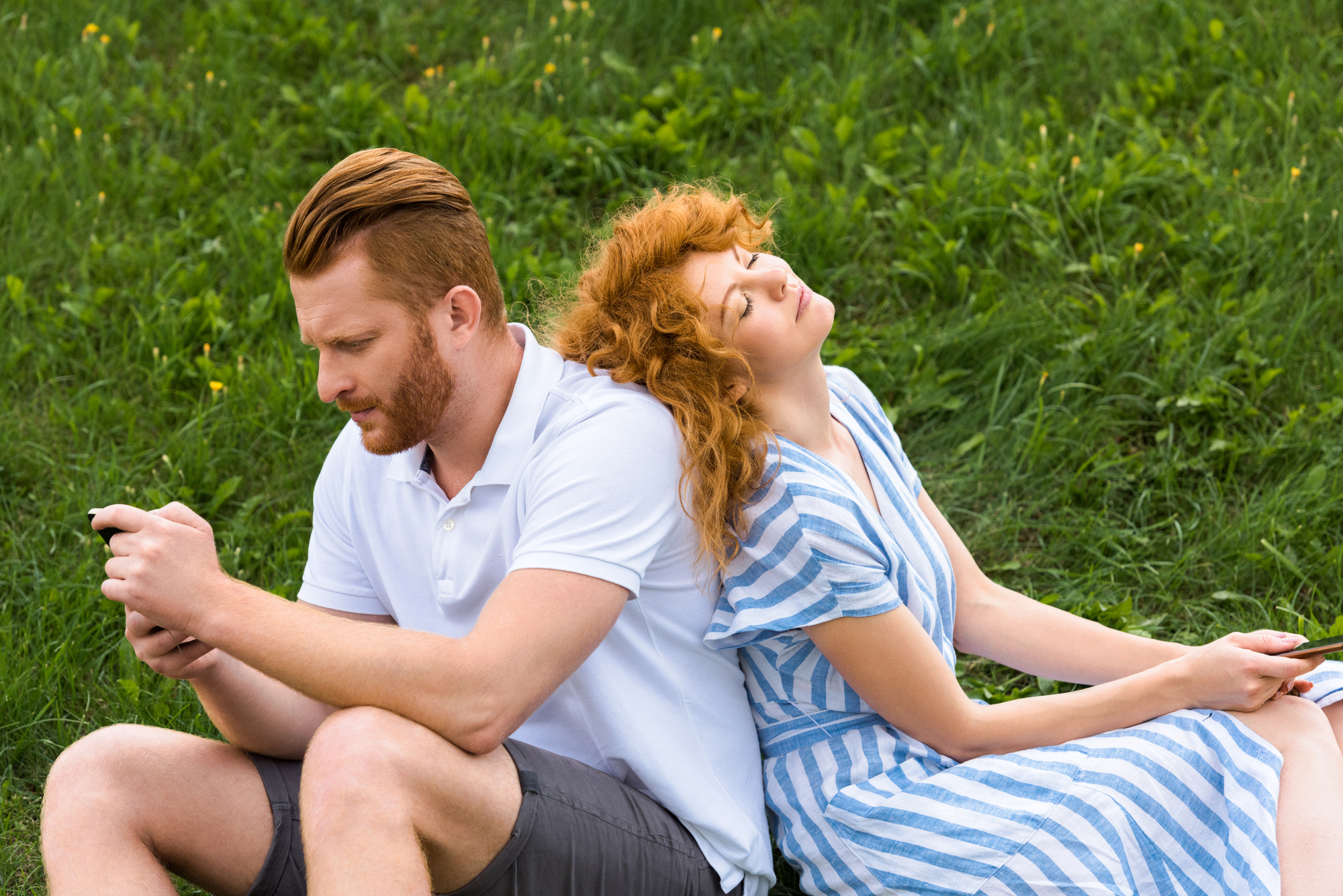 A man and a woman are sitting back-to-back on a grassy field. The man, with a serious expression, is focused on his smartphone, while the woman, wearing a striped dress, leans her head back with her eyes closed, looking relaxed.