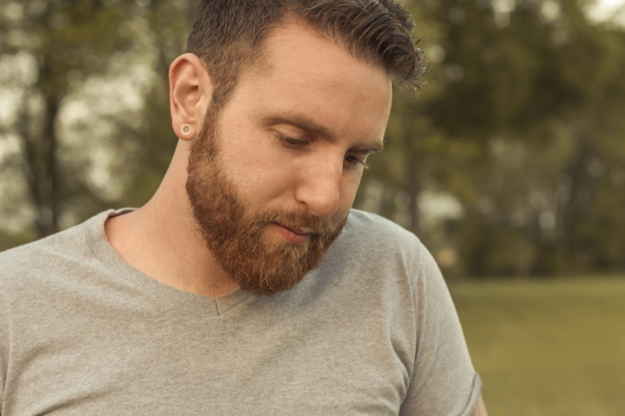 A man with a beard, short hair, and ear gauges is looking down. He is wearing a light gray T-shirt and standing outdoors on a green grassy area, with trees visible in the blurred background. The lighting suggests it's a calm, possibly cloudy day.