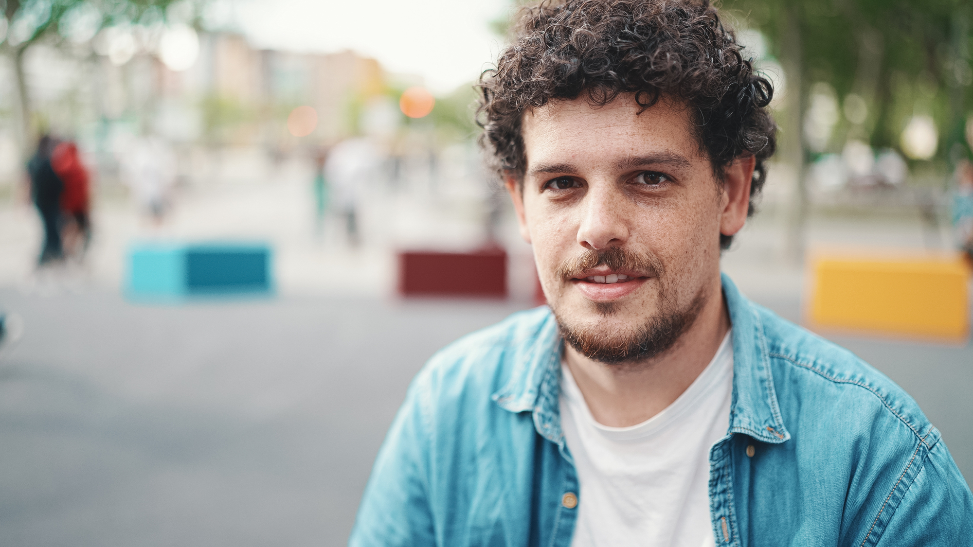 A man with curly hair and a beard is smiling slightly while looking at the camera. He is wearing a white t-shirt and a denim shirt. The background is an outdoor setting with blurred trees and colorful rectangular blocks.