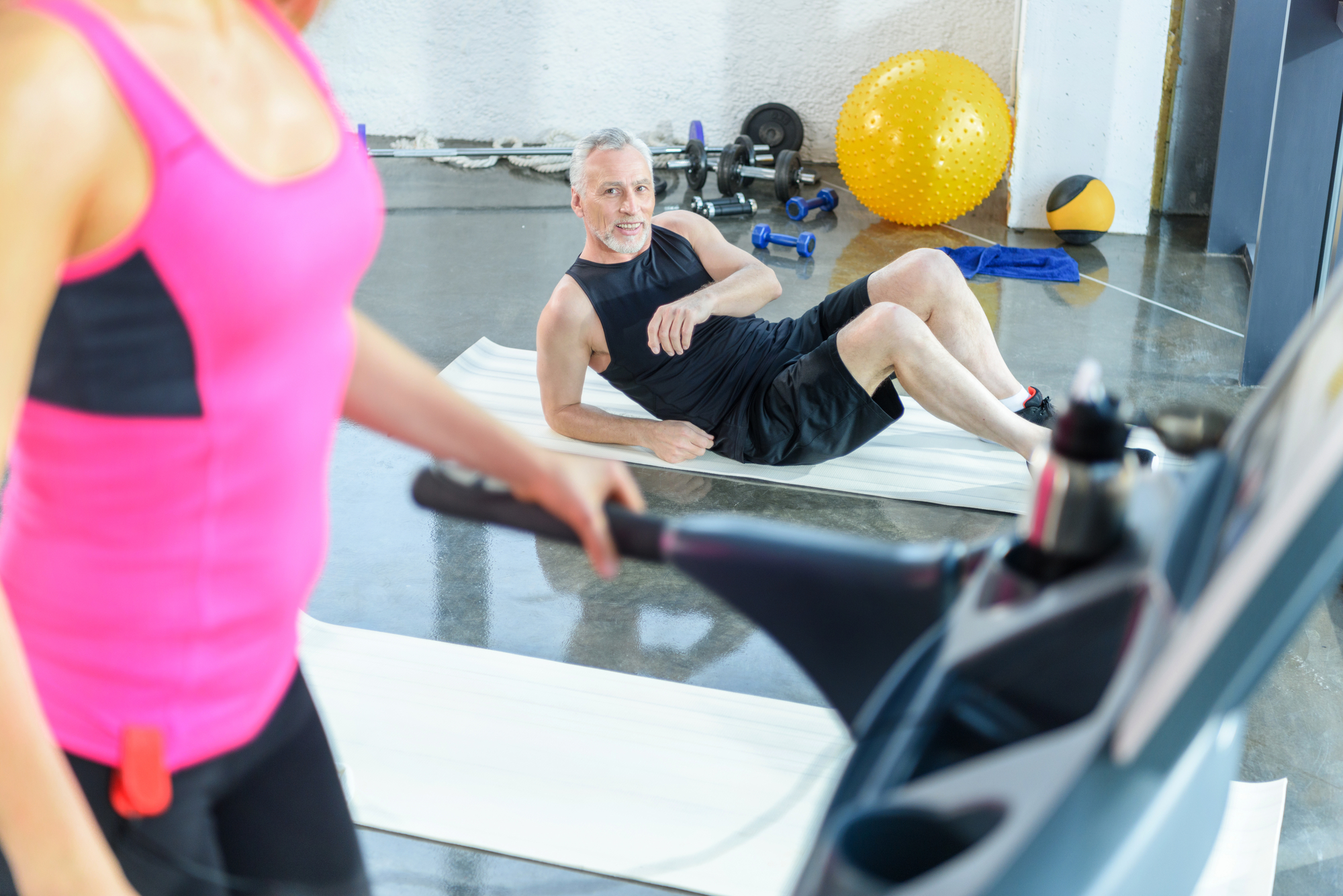 A man with gray hair and a black workout outfit is performing a side plank on a white exercise mat in a gym. Nearby, a woman in a pink top is on a treadmill, partially visible. Gym equipment like dumbbells and a yellow exercise ball are in the background.
