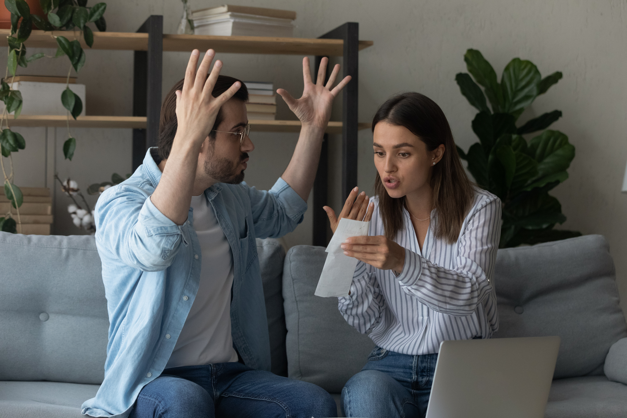 A man and woman are sitting on a couch in a living room, engaged in a heated discussion. The man has his hands raised in frustration, while the woman is holding a piece of paper and gesturing as she talks. Shelves with books and plants are in the background.