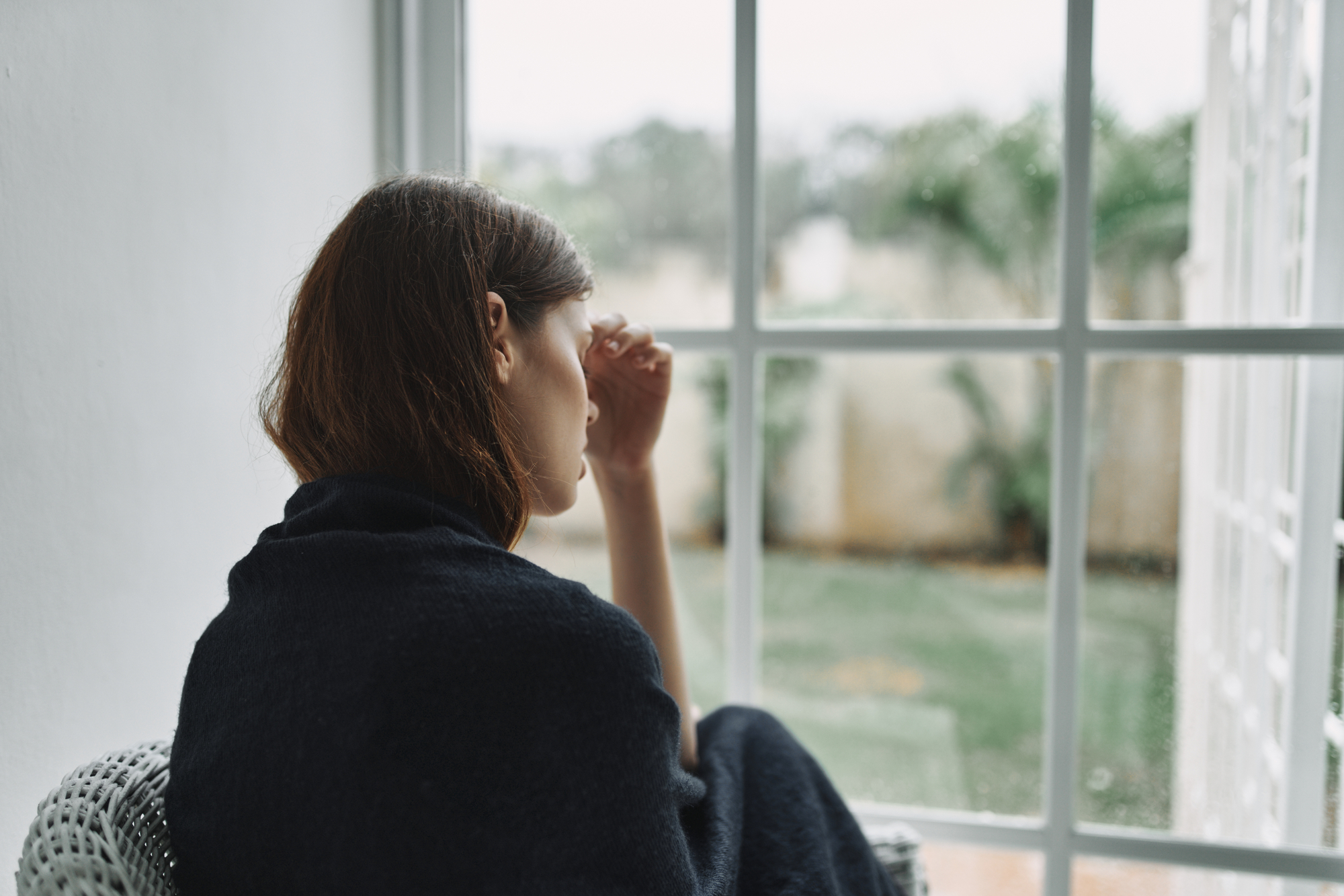 A woman with shoulder-length brown hair wrapped in a dark blanket is sitting on a white wicker chair indoors, looking out a window. Her hand rests on her forehead, and she appears to be in thought, gazing out at a rainy outdoor scene with greenery in the background.