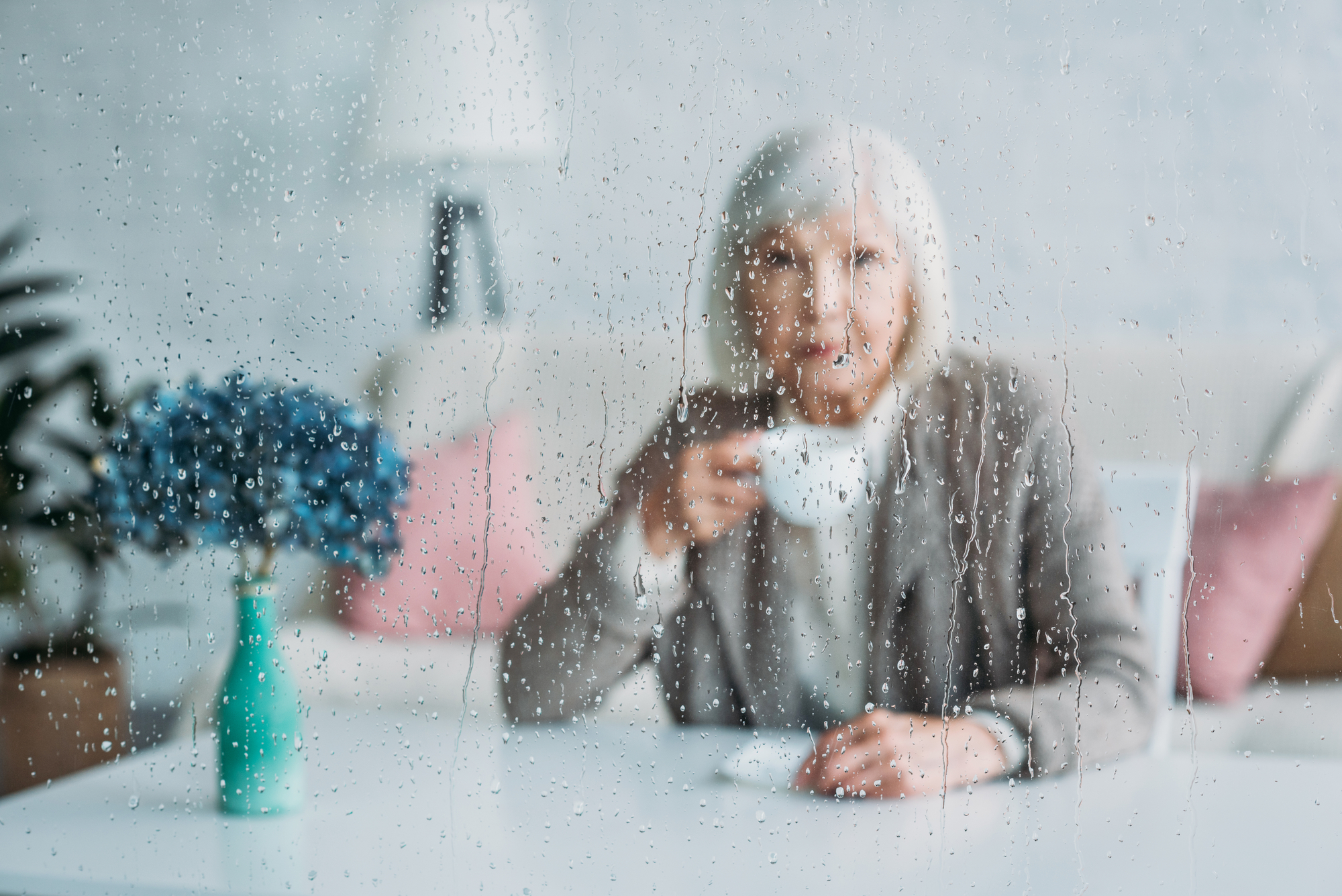 An elderly woman with white hair is sitting at a table indoors, holding a cup of coffee. The image is taken through a rain-soaked window, with raindrops clearly visible. A green vase with blue flowers is on the table next to her.