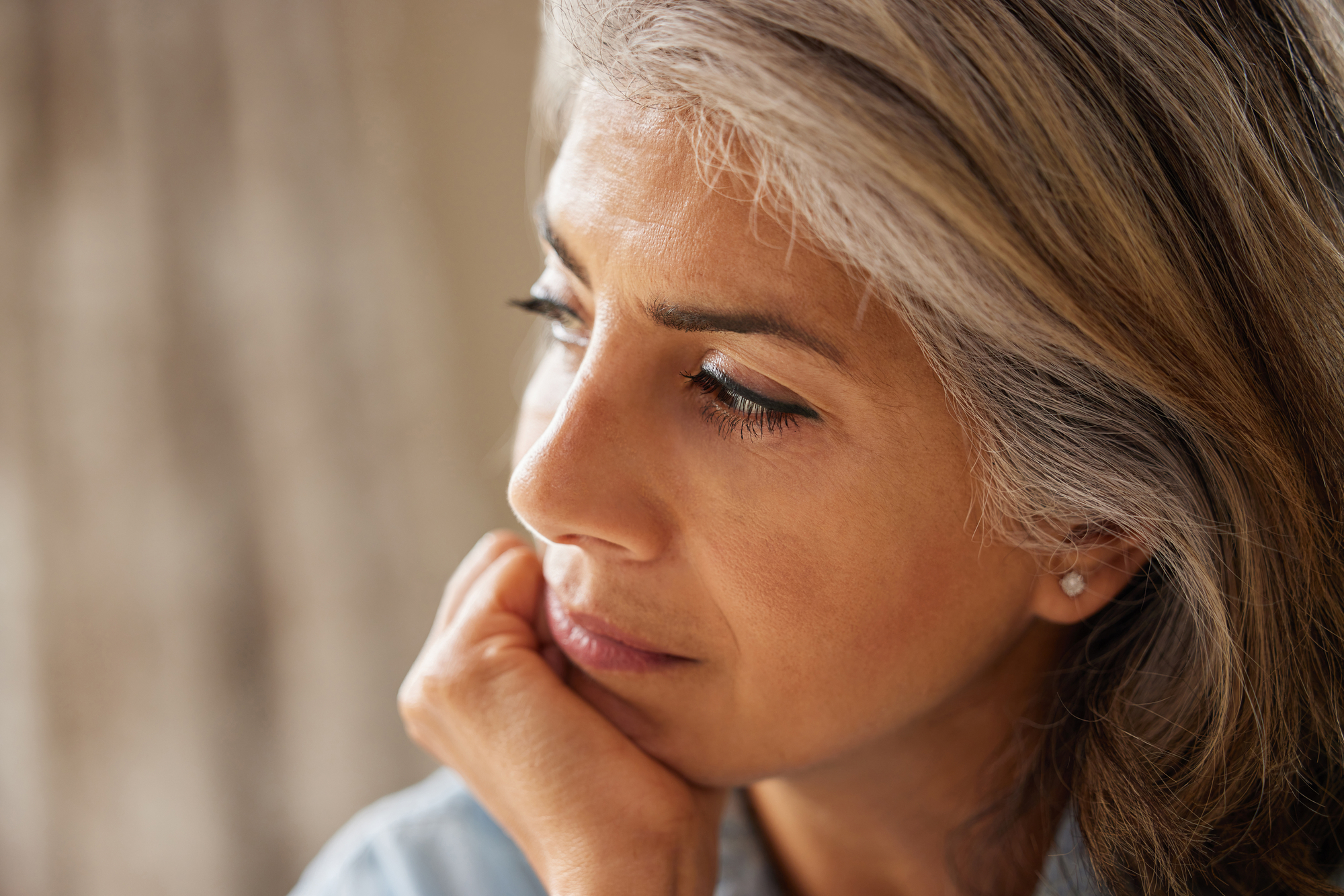 Close-up of a woman with wavy gray and brown hair, resting her chin on her hand. She has a contemplative expression and is looking off to the side. The background is blurred, focusing attention on her thoughtful face.